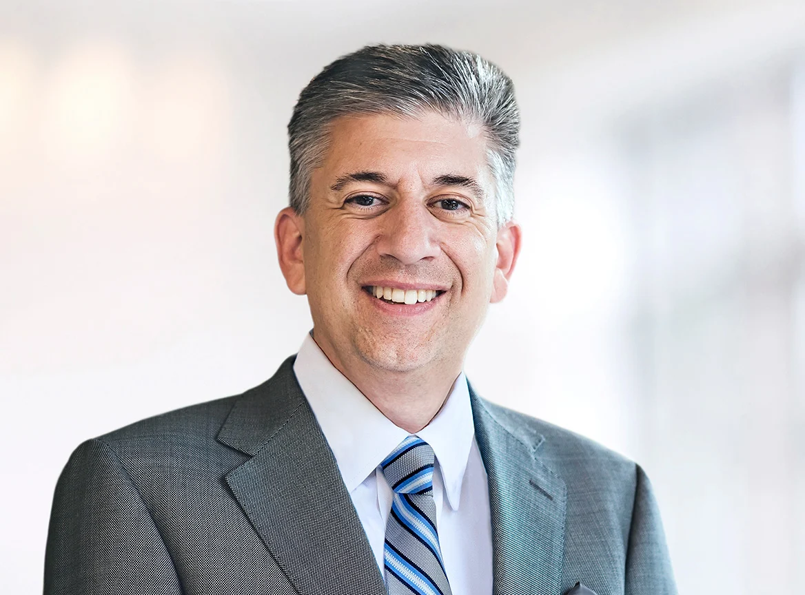 Professional headshot of a middle-aged man with short salt-and-pepper hair, smiling broadly, wearing a gray suit with white shirt and blue striped tie