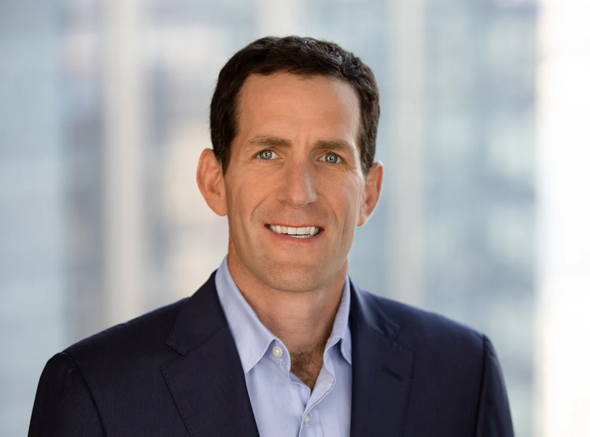 Professional headshot of a man with short dark hair and blue eyes, smiling, wearing a navy blazer and light blue shirt, against a blurred office background with windows.