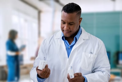 A person in a lab coat holding two small boxes, with blurred background figures.