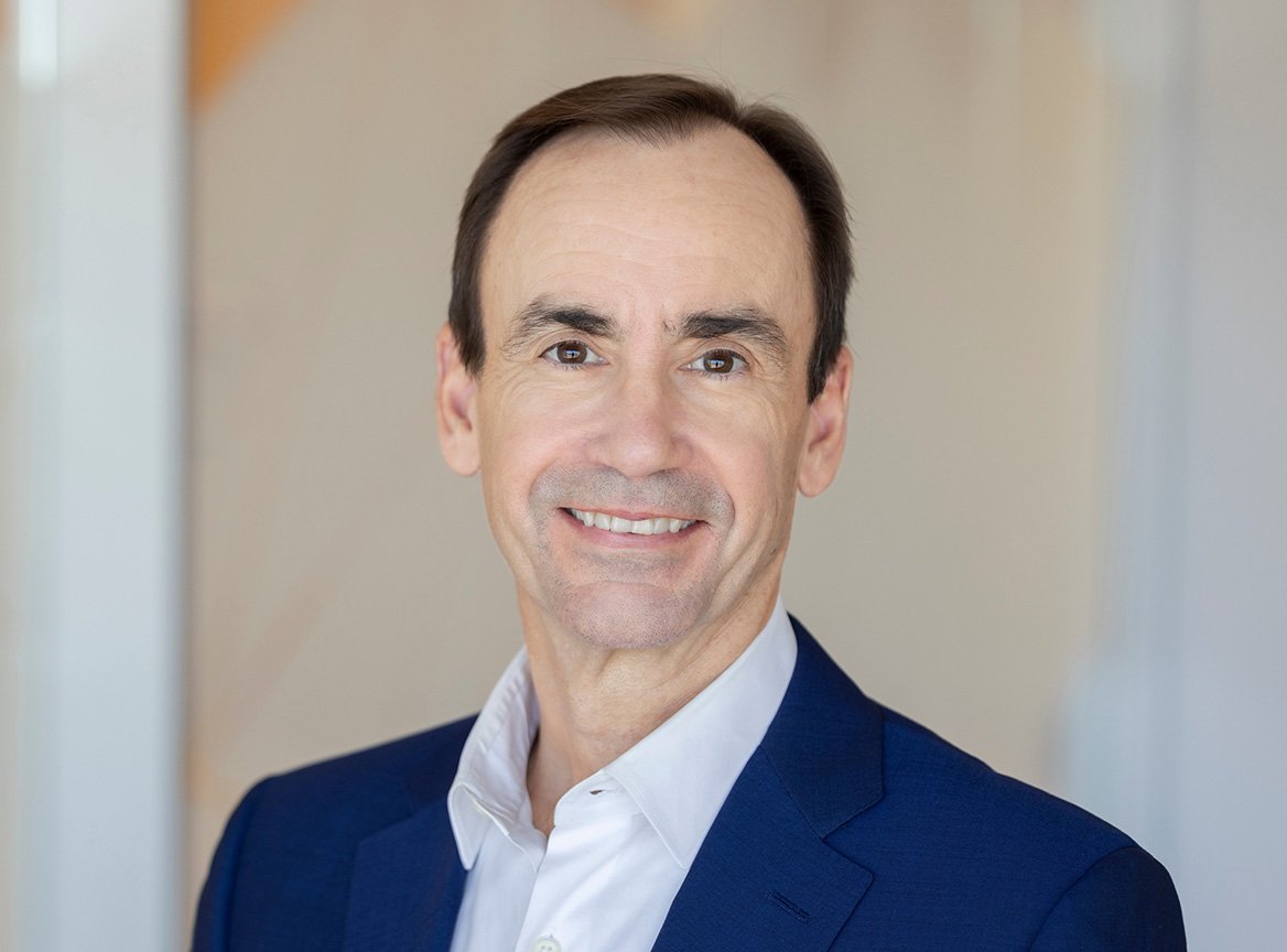 Professional headshot of a middle-aged man with short dark hair, smiling, wearing a navy blue blazer and white button-up shirt against a light background