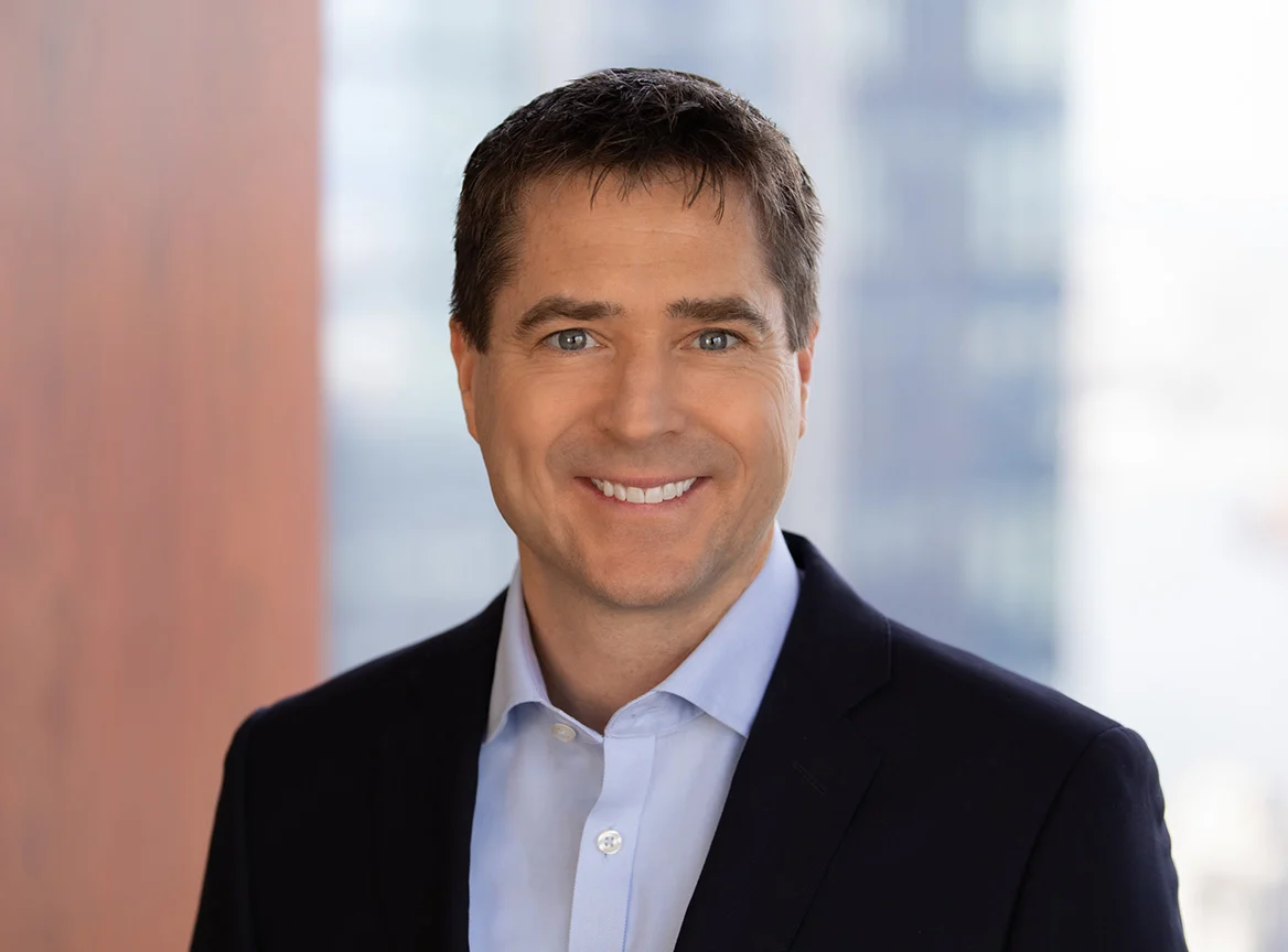 Professional headshot of a middle-aged man with short brown hair and blue eyes, smiling confidently, wearing a navy blue blazer with light blue button-up shirt, against a softly blurred office background.