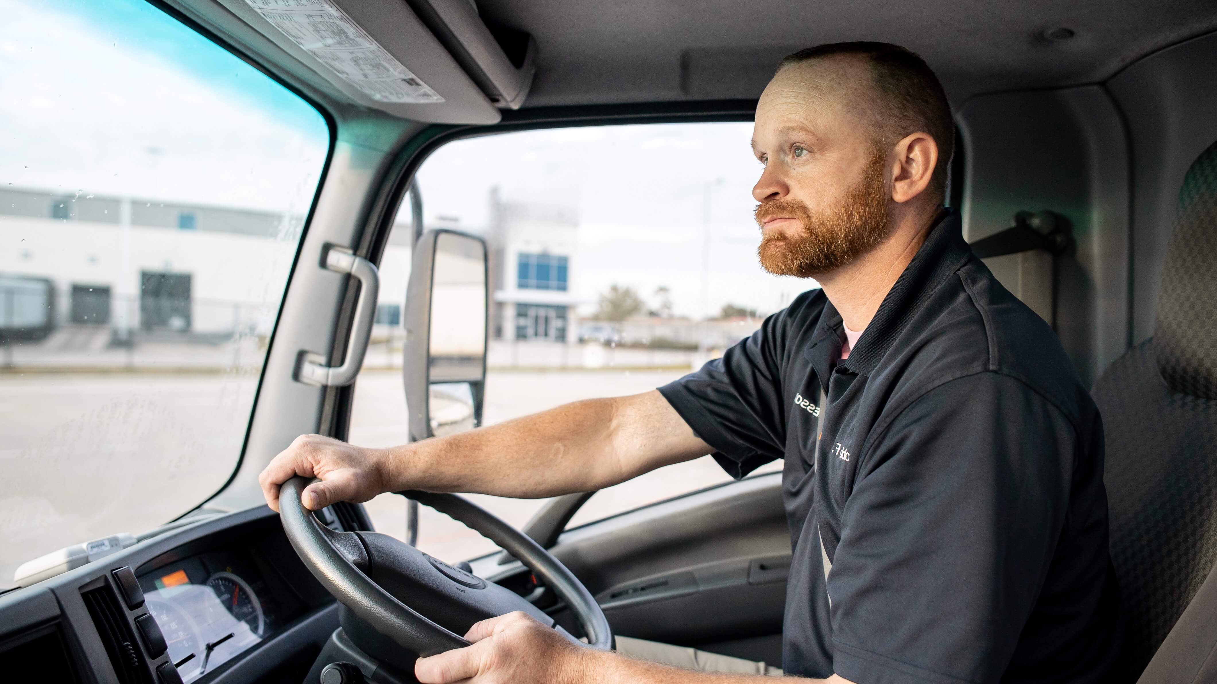 Truck driver steering a vehicle, wearing a black polo, with an industrial building visible through the window.
