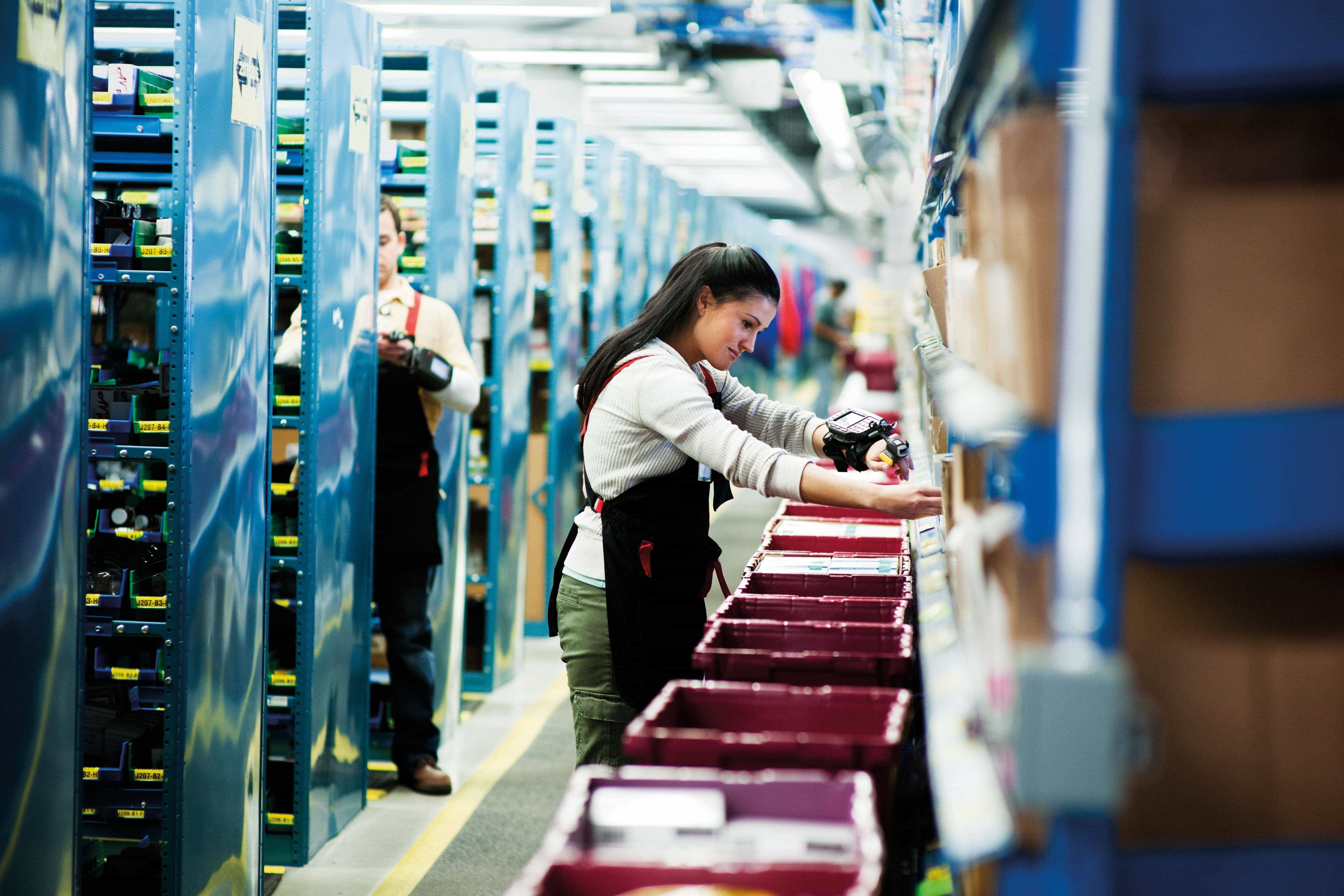 Warehouse workers sorting and scanning items on shelves with red bins in an organized storage facility.