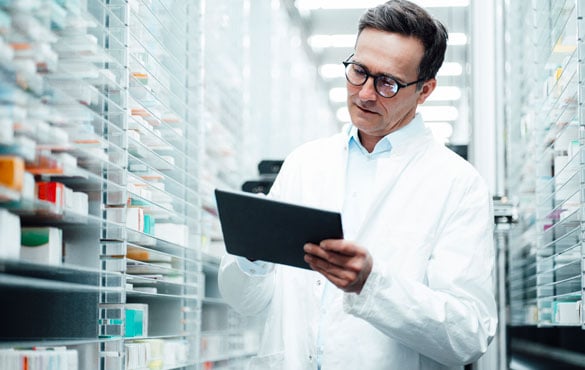 A pharmacist in a white lab coat holds a clipboard, standing between shelves of medication.