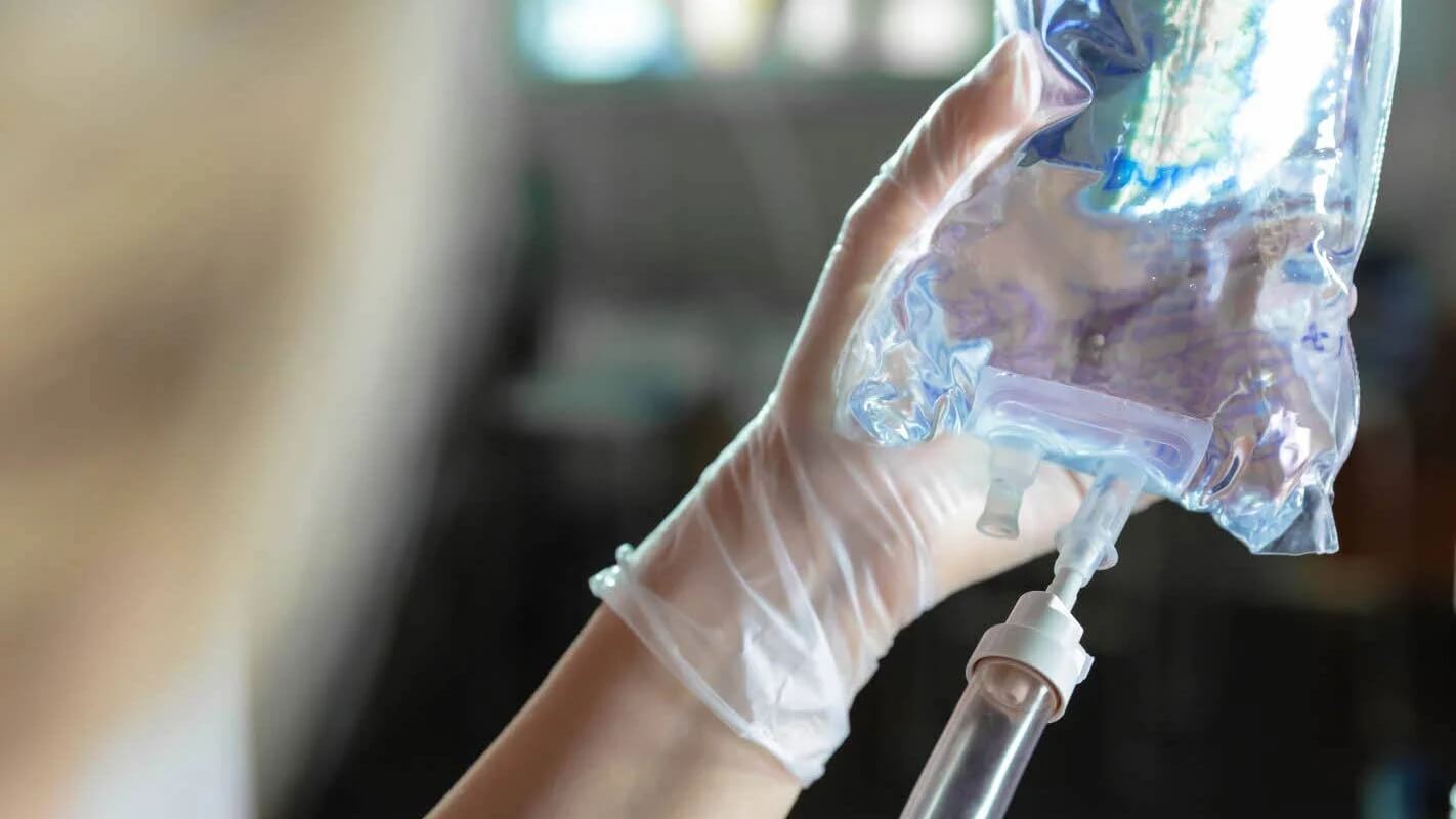 A gloved hand holds an IV fluid bag with a tube attached, preparing for medical use.