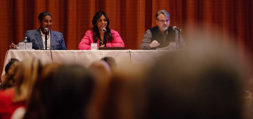 Three people sit at a panel table with microphones in front of an audience.
