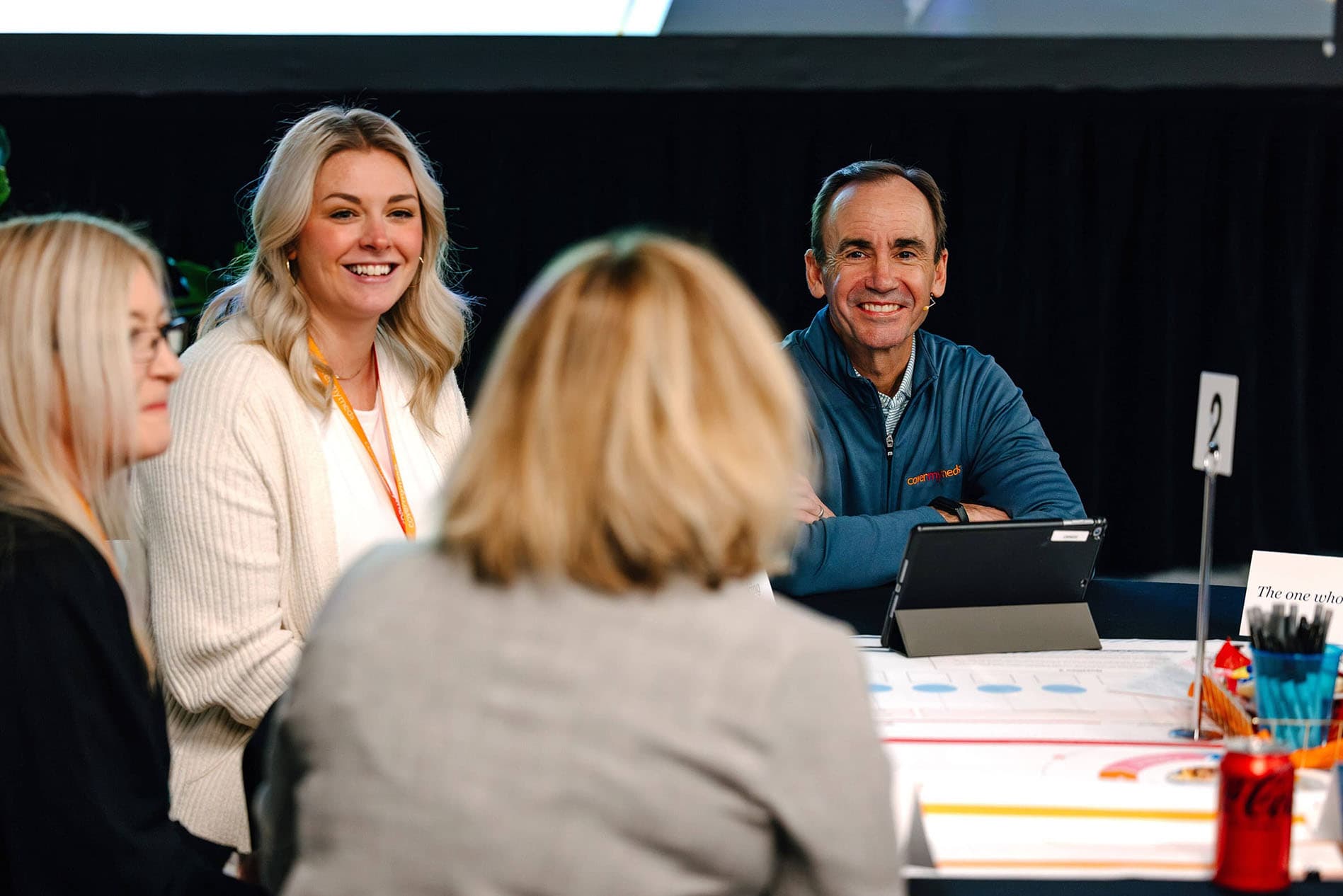 Four people sitting around a table with papers and a tablet, engaged in discussion. A sign with the number 2 is on the table.