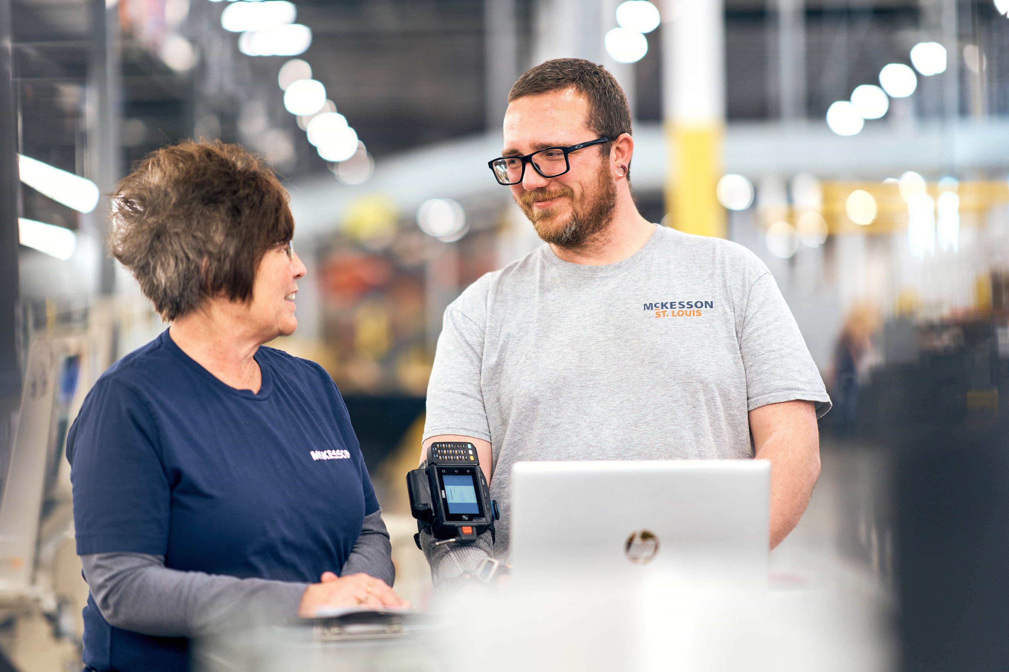 Two people in a warehouse setting, one holding a device and the other looking at them. Both wear shirts with "McKesson" logos.