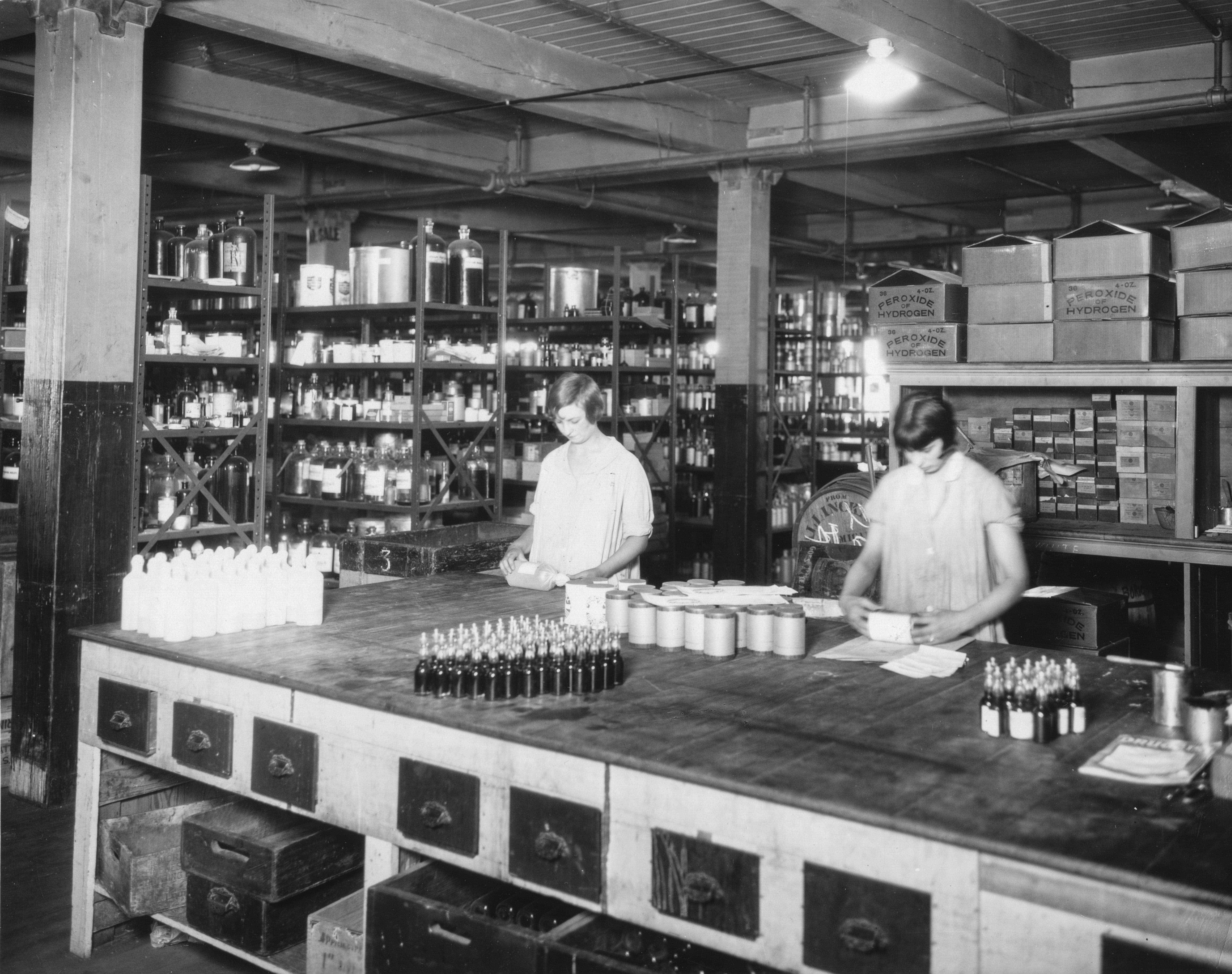 A historical black and white photo of two people working at a large table in a warehouse filled with shelves of various bottles and boxes.