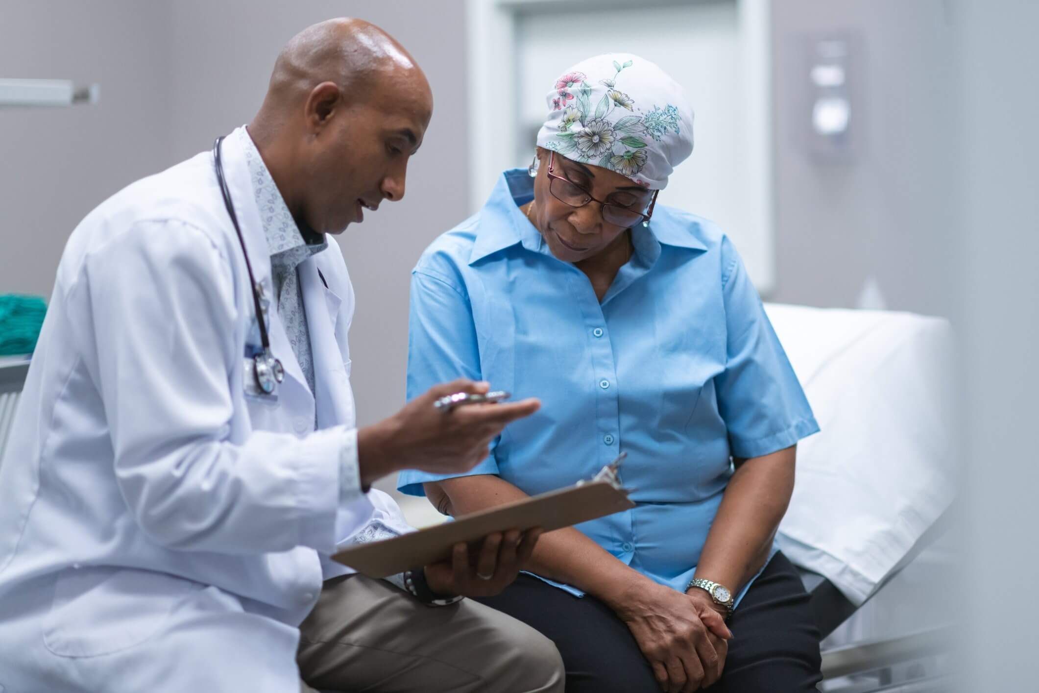 A doctor in a white coat consults with a patient wearing a headscarf and blue shirt in a medical examination room, holding a clipboard.