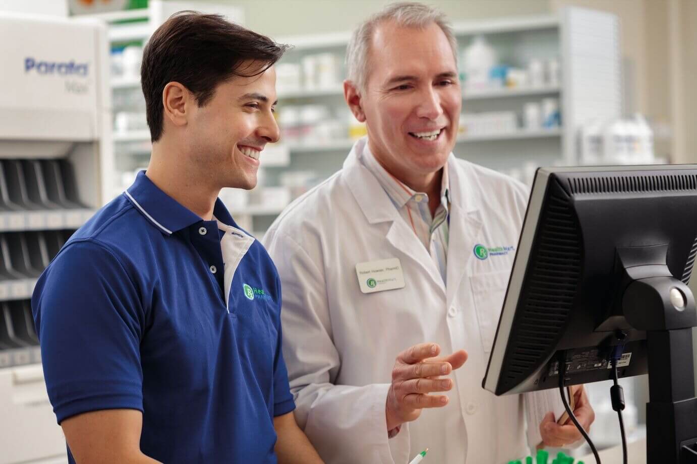 Two pharmacists, one in a blue polo shirt and the other in a white lab coat, discuss something while looking at a computer screen in a pharmacy.