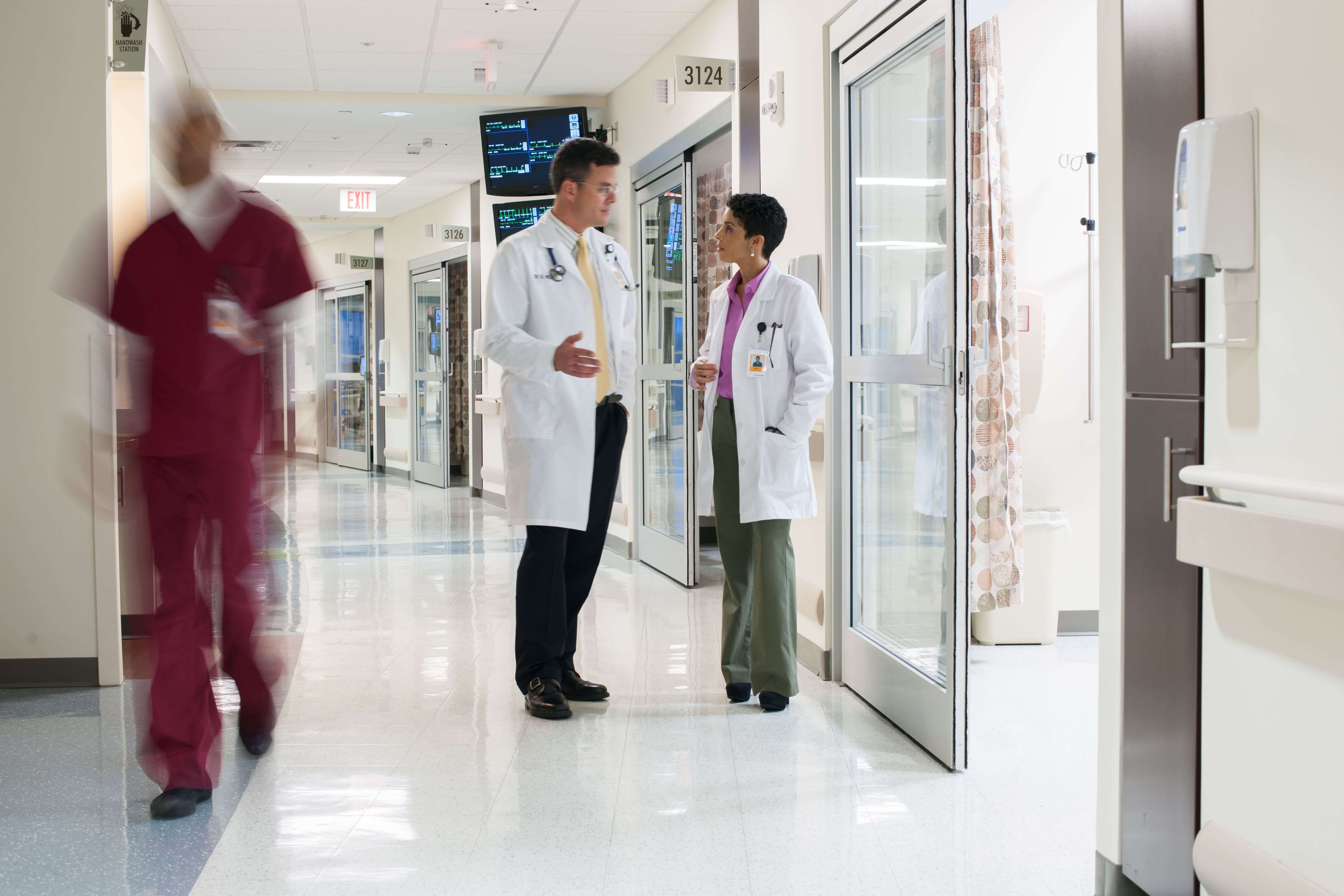 Two doctors in white coats converse in a hospital hallway while a nurse in red scrubs walks by.