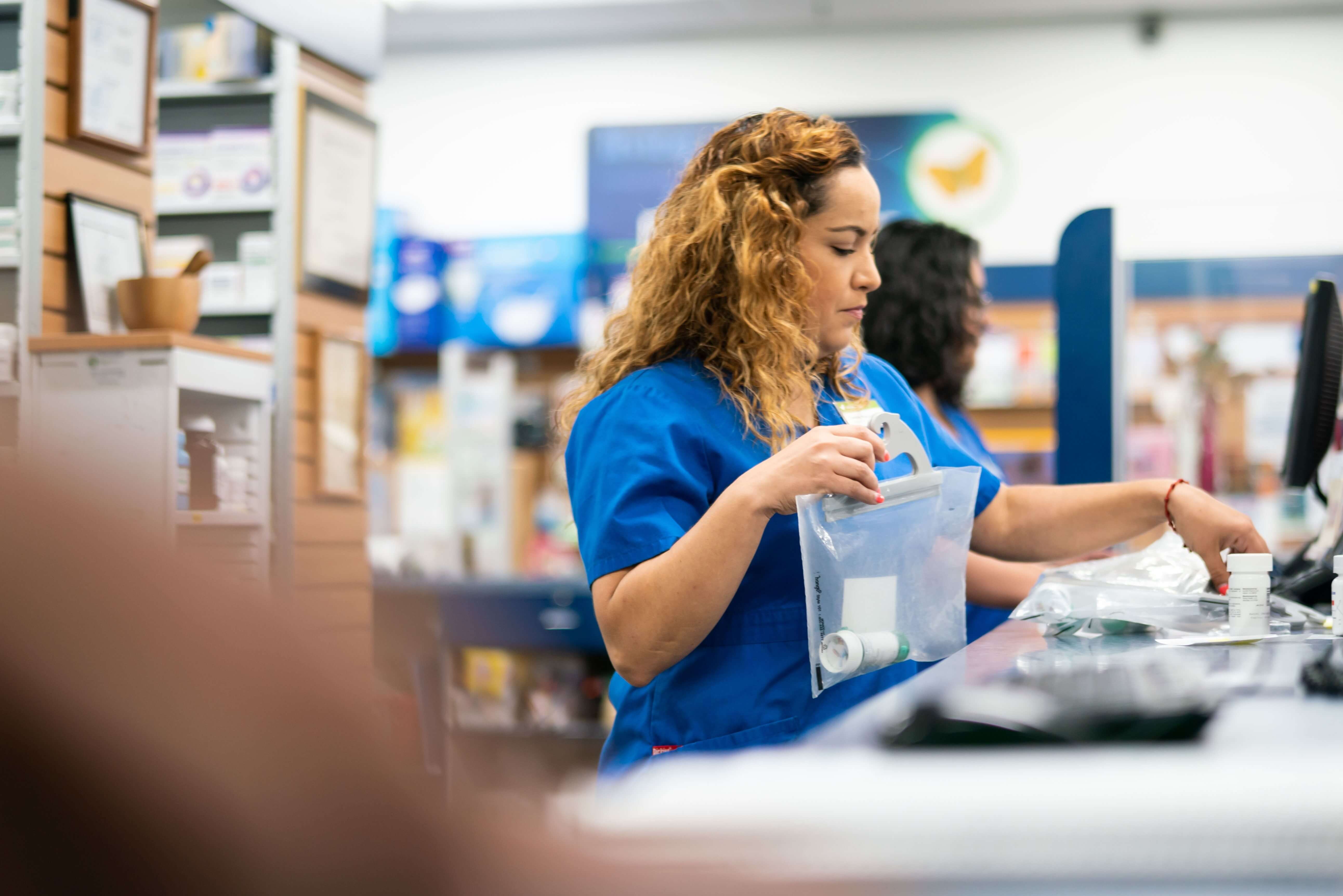 A pharmacy worker in a blue uniform holds a clear plastic bag with medication bottles at a counter.