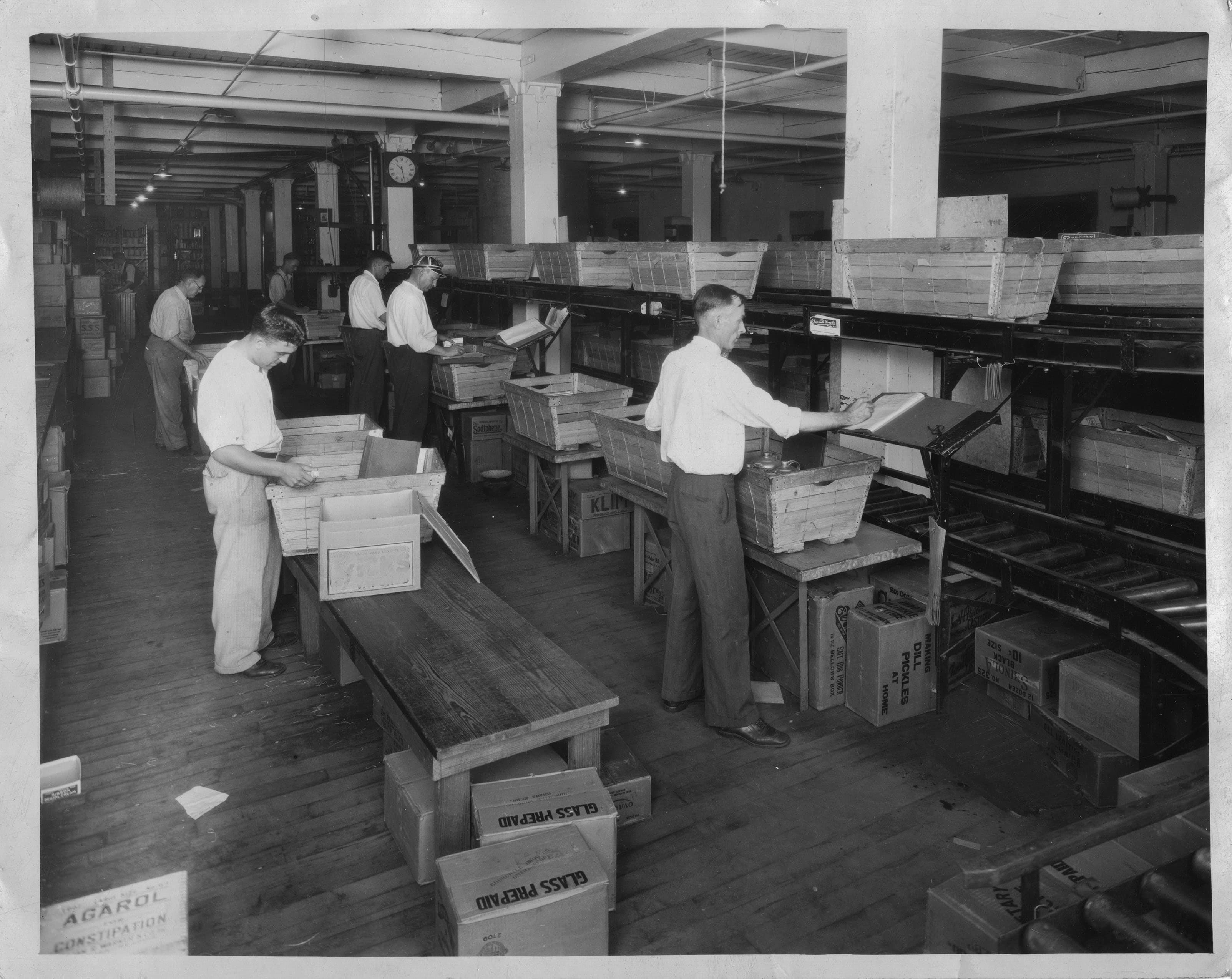 A historical black and white image of men working in a factory, sorting and packing items into boxes on a conveyor belt.