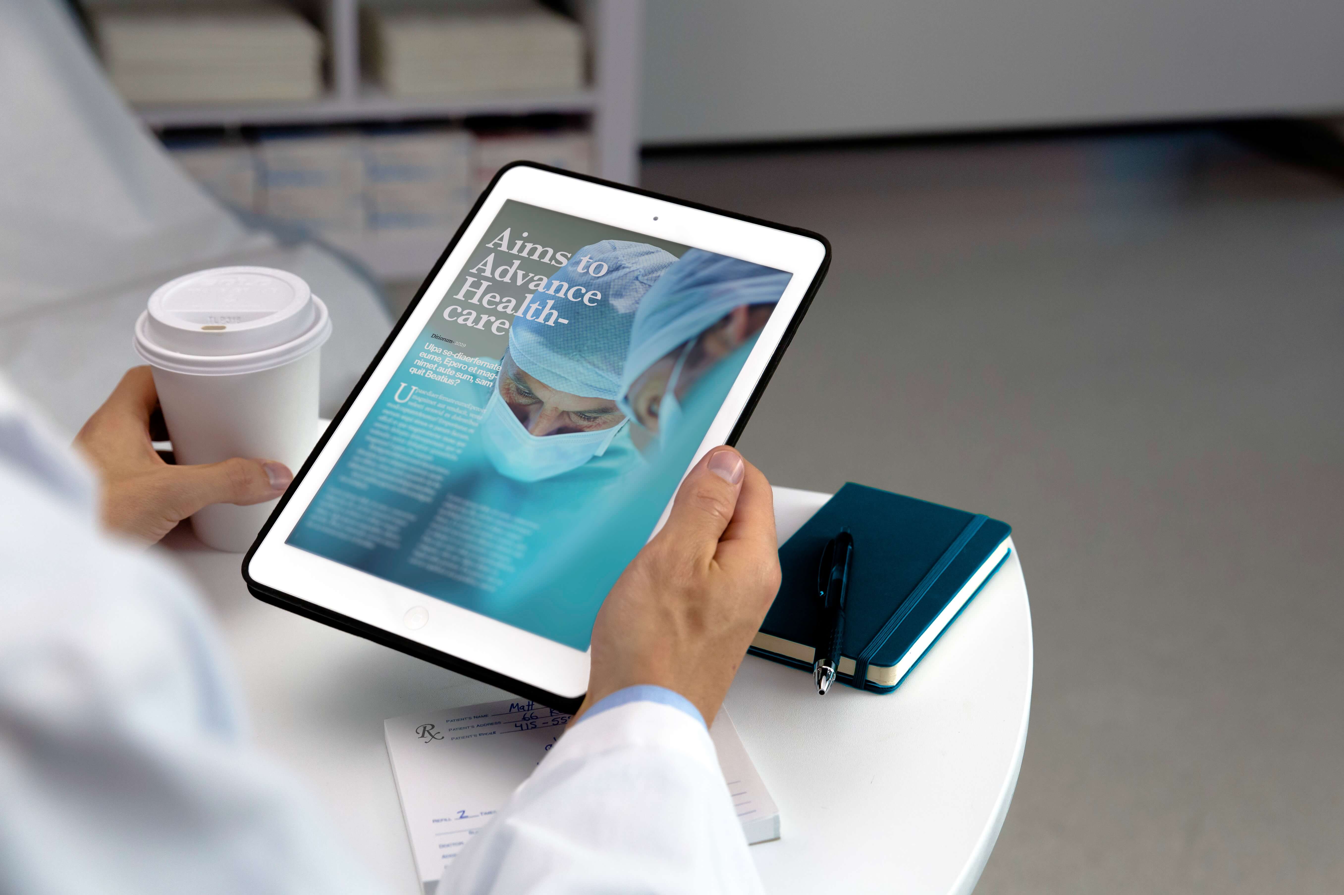 A person in a white coat holds a tablet displaying a healthcare article, with a coffee cup and notebook nearby.