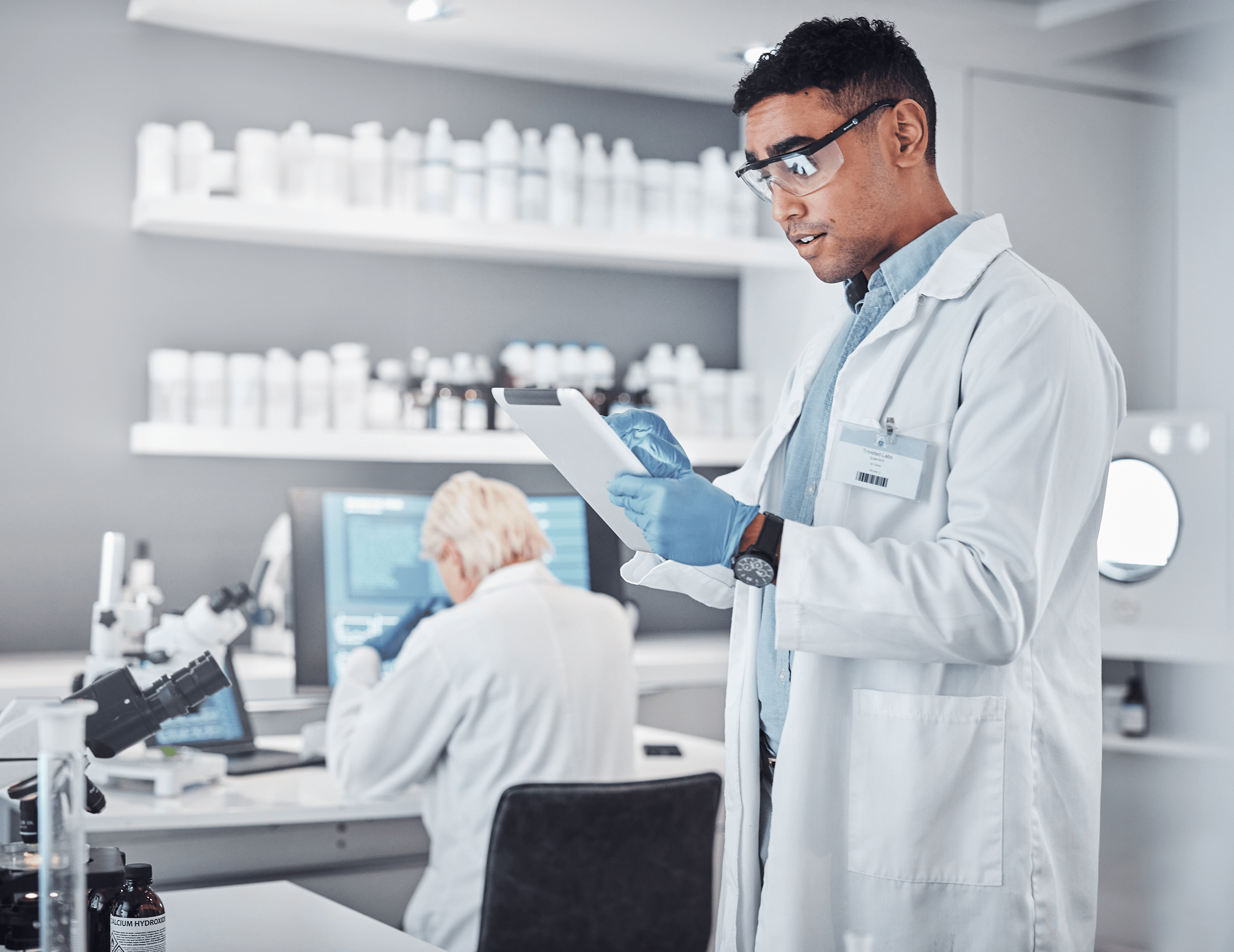 Two scientists in a laboratory, one standing and reading a tablet, the other sitting and working at a computer.