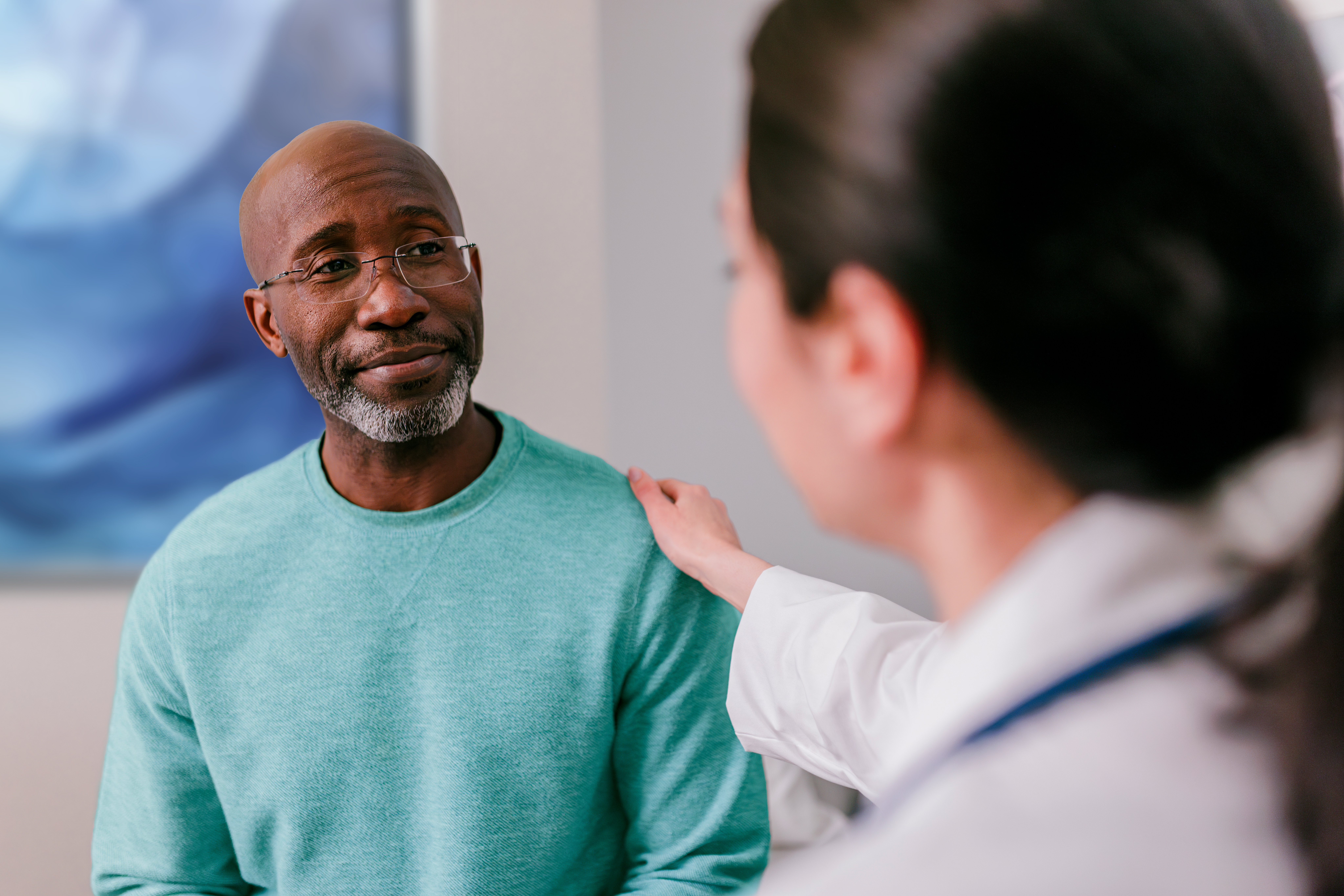 A doctor in a white coat places a reassuring hand on the shoulder of a patient wearing a green sweater.