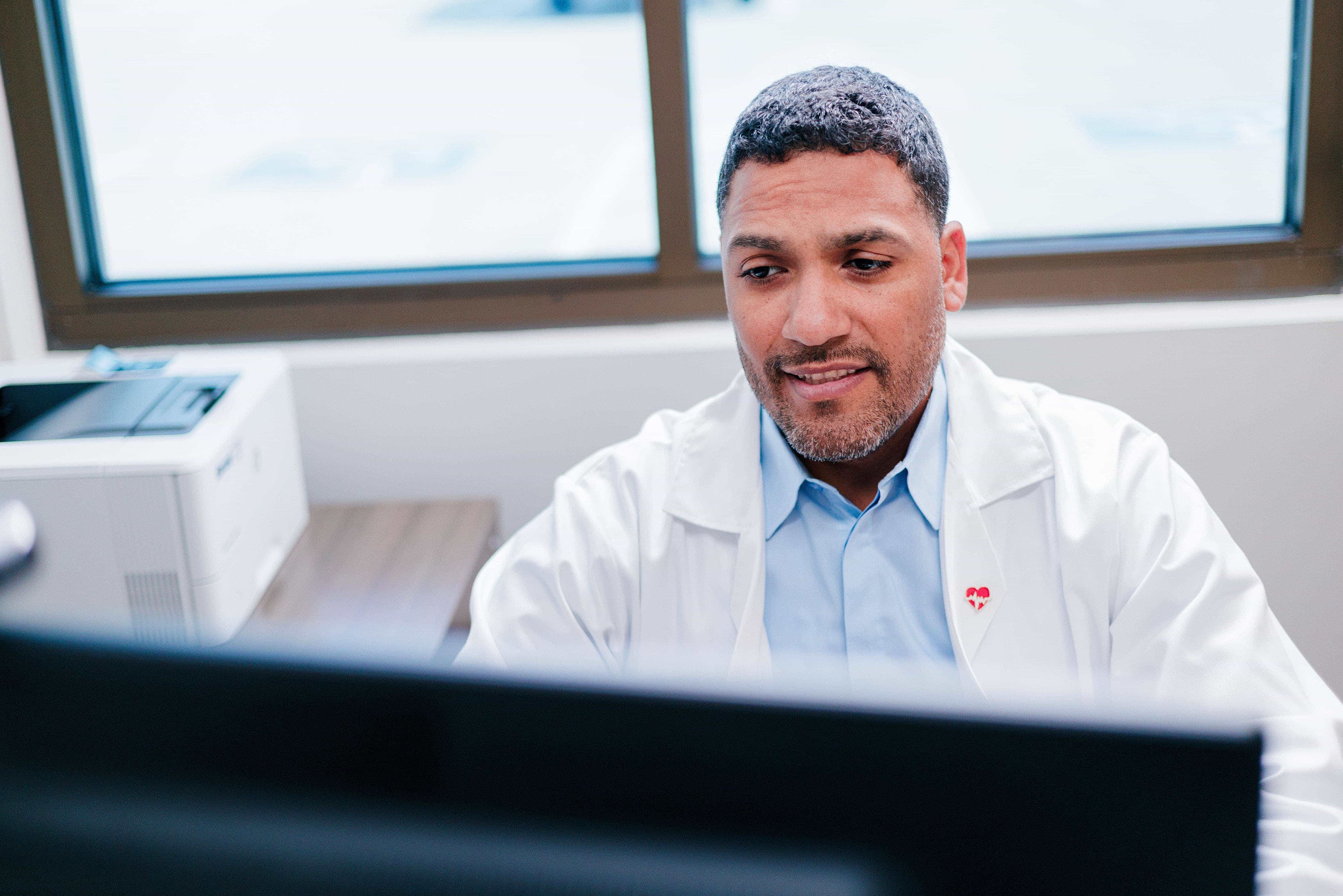 A person in a white coat works at a computer in an office with a printer on the desk.