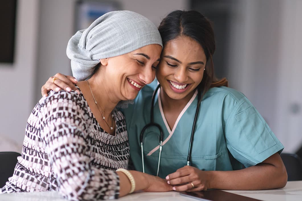 A healthcare worker in scrubs and a stethoscope comforts a patient wearing a headscarf