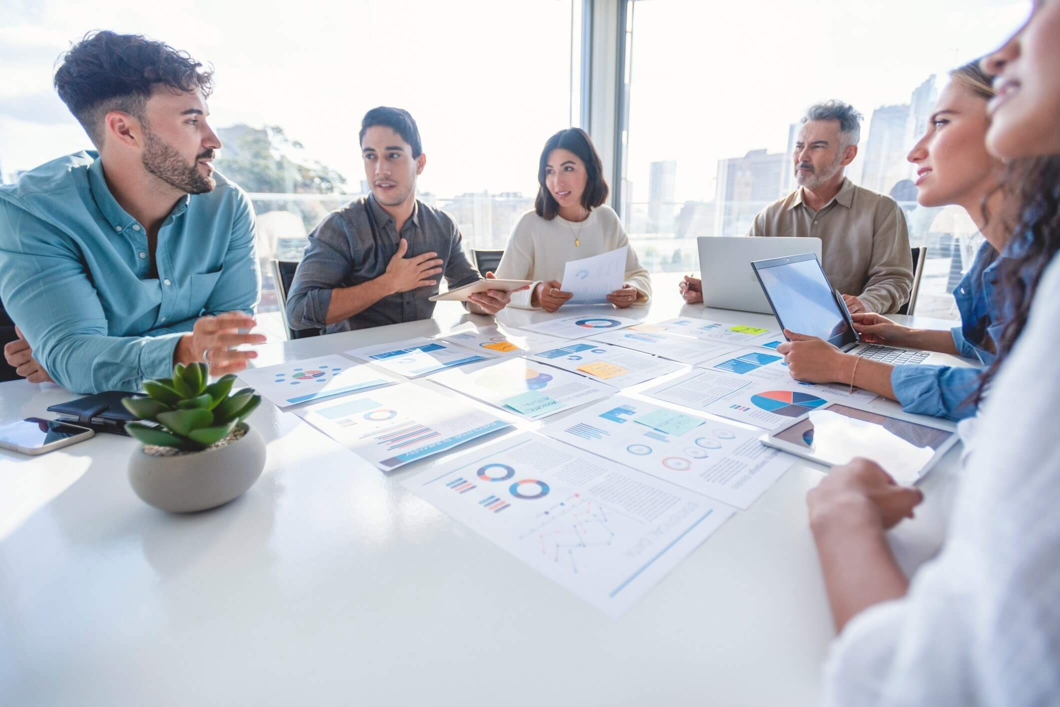 Six people in a meeting room discussing documents with charts and graphs spread on the table.
