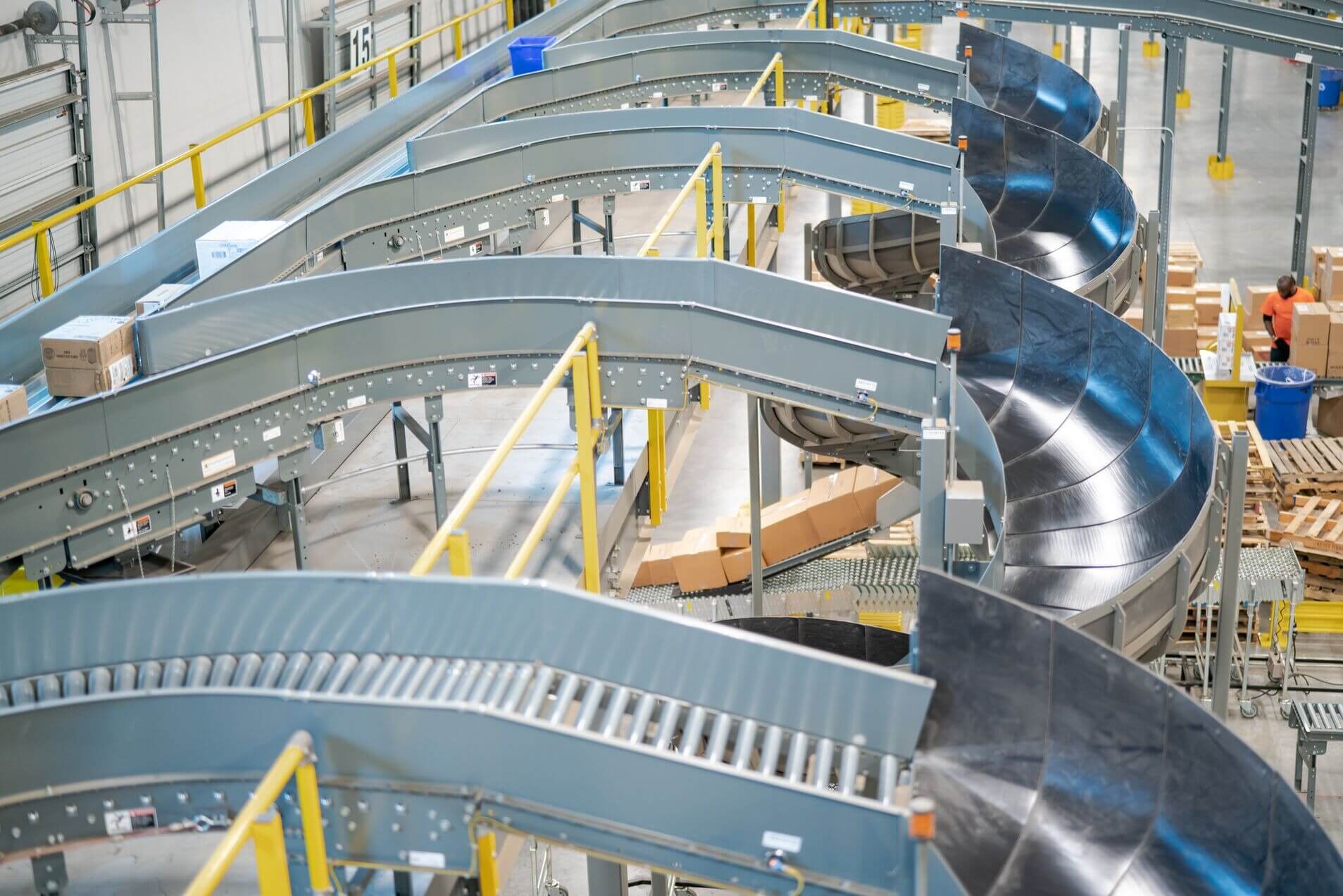 Warehouse with multiple conveyor belts transporting packages, including curved and spiral sections. Workers are visible in the background.