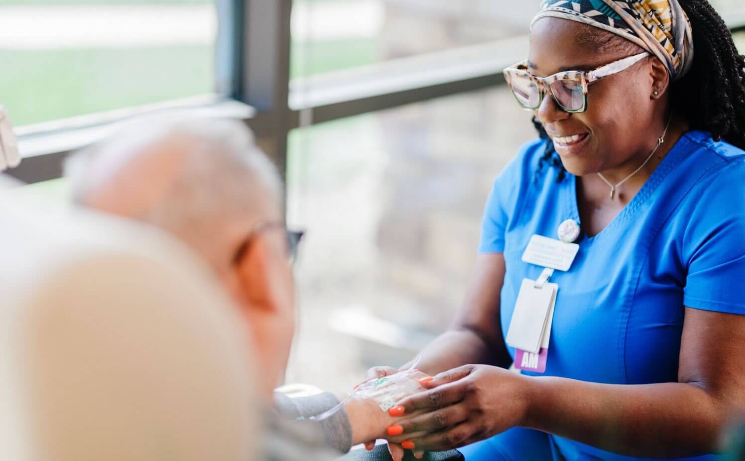 Healthcare worker in blue scrubs holding the hand of a patient near a window.