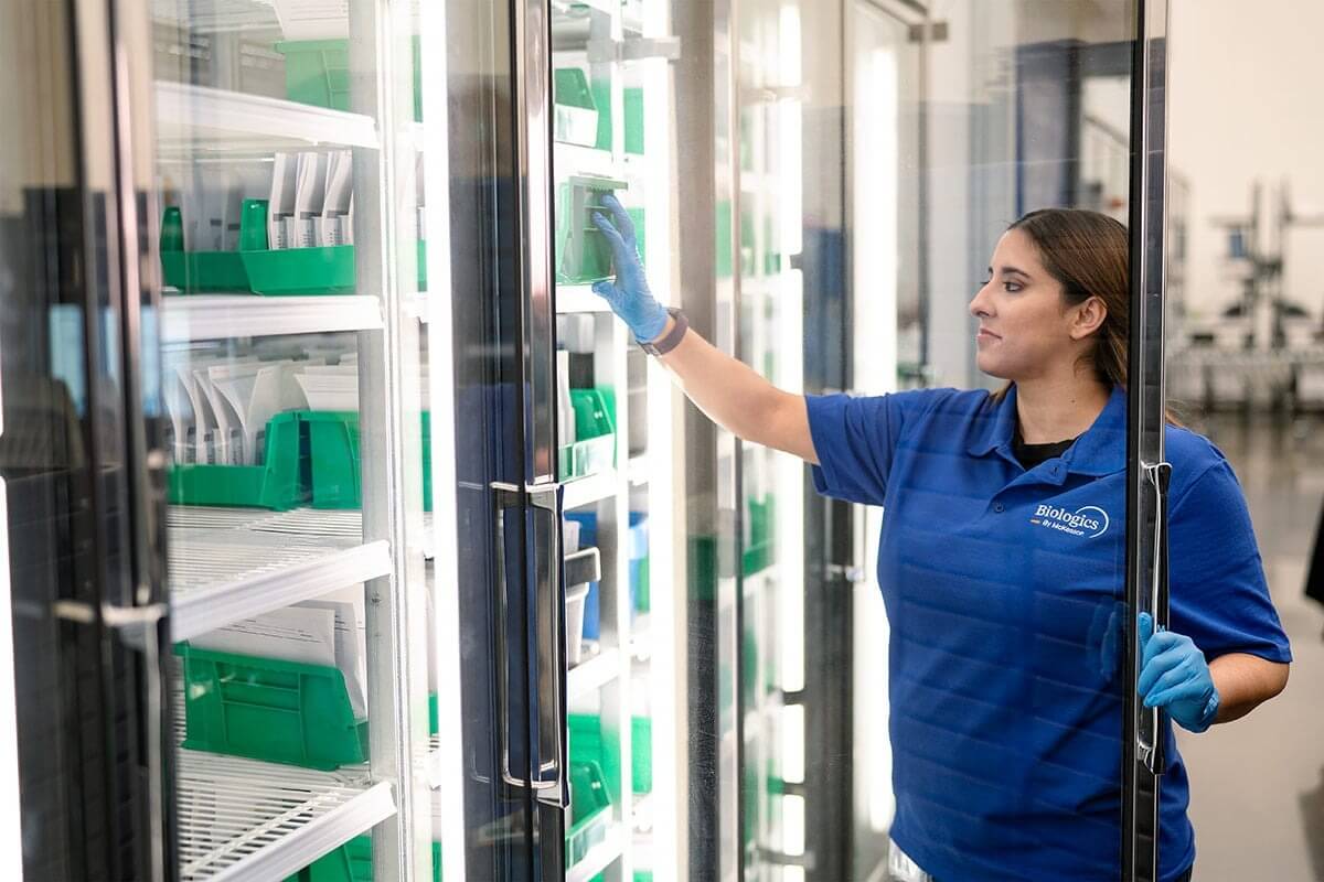 Person in blue uniform and gloves accessing items from a refrigerated storage unit with green and white containers.
