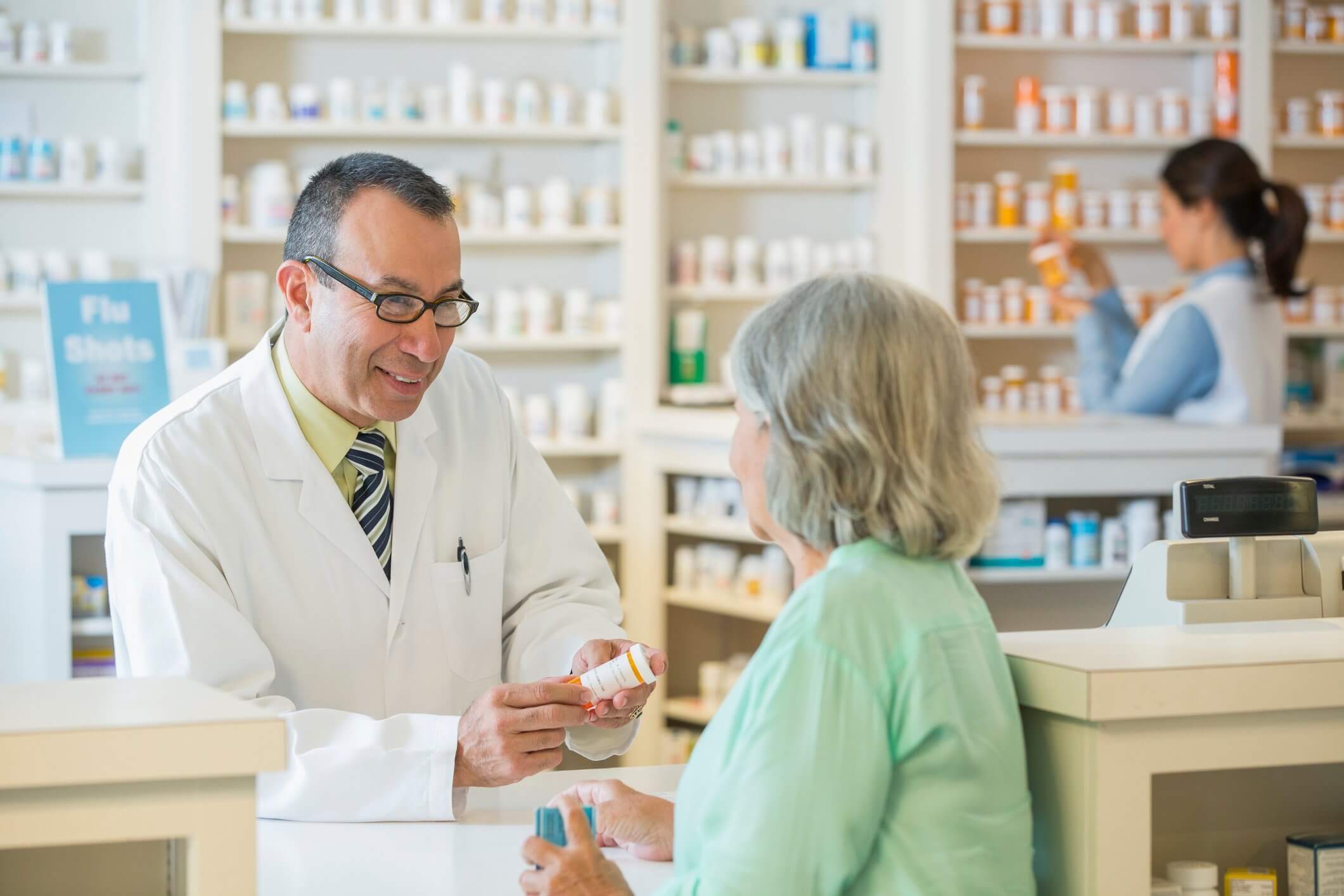 Pharmacist in a white coat discussing medication with an elderly woman at a pharmacy counter, while another person organizes shelves in the background.