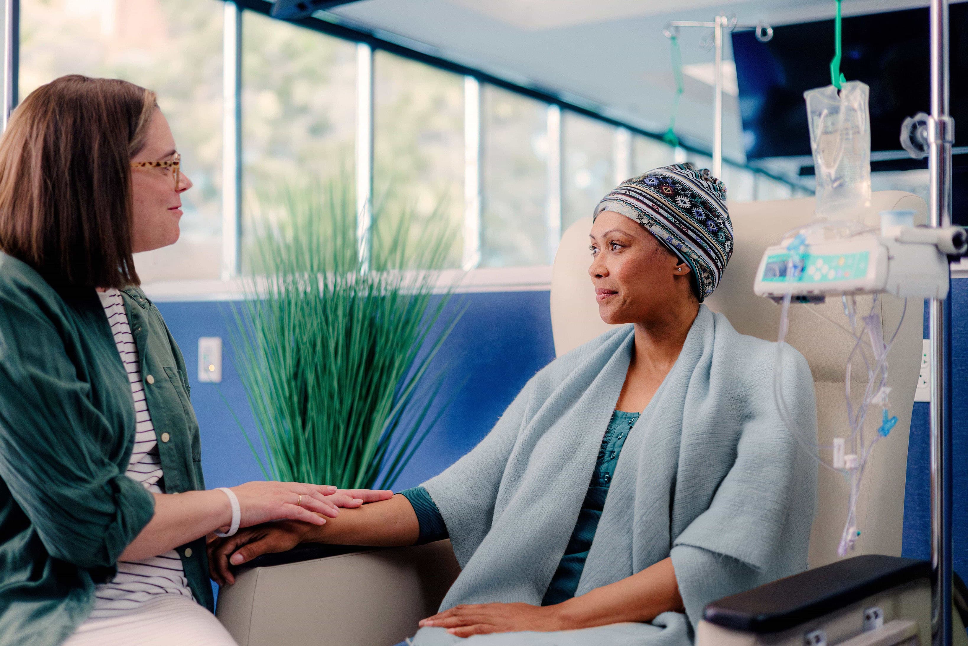 Two people in a medical setting, one sitting in a chair receiving IV treatment, the other sitting beside them holding their hand.