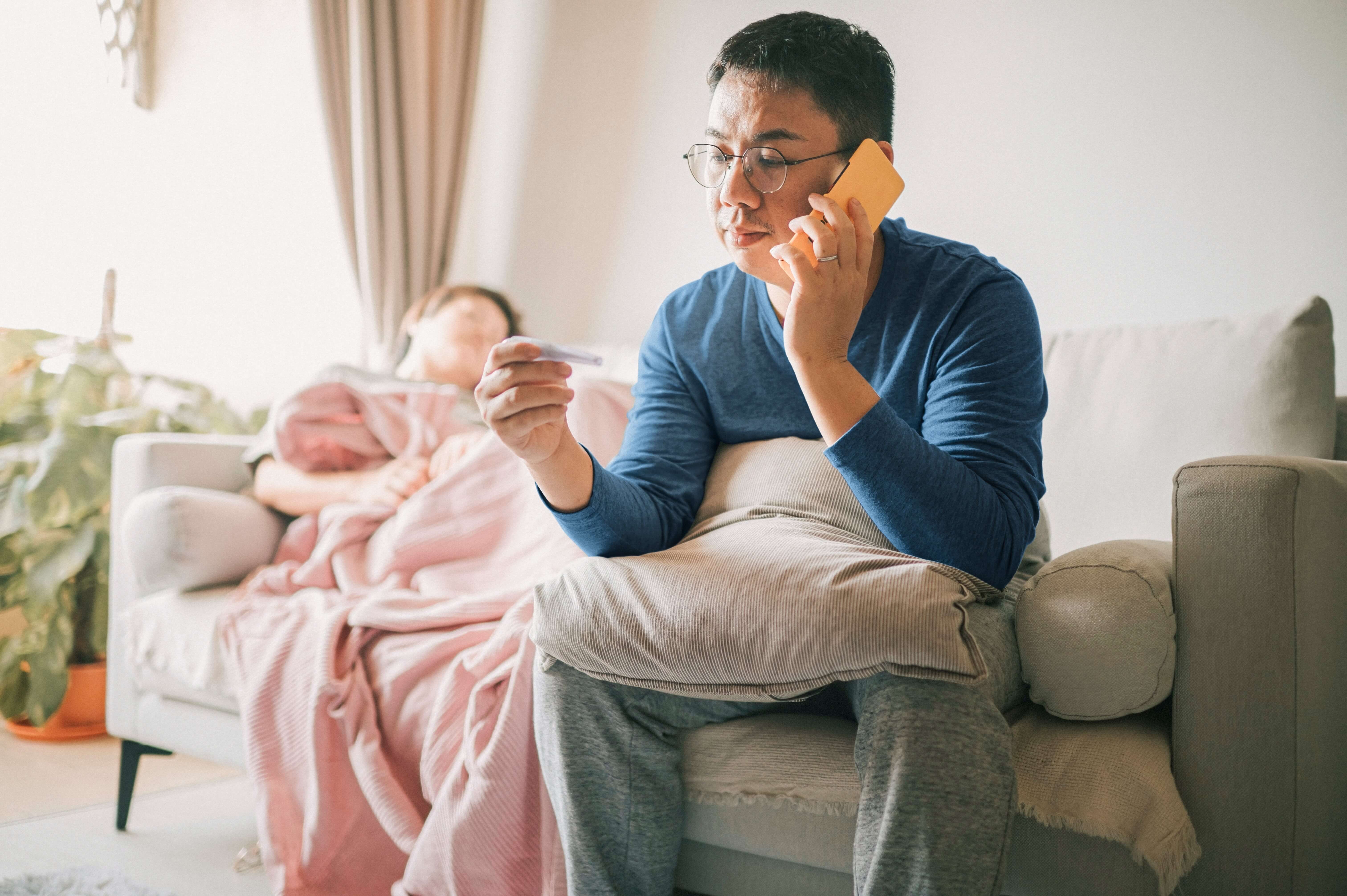 A person in a blue shirt sits on a couch holding a credit card and phone, while another person sleeps under a pink blanket in the background.