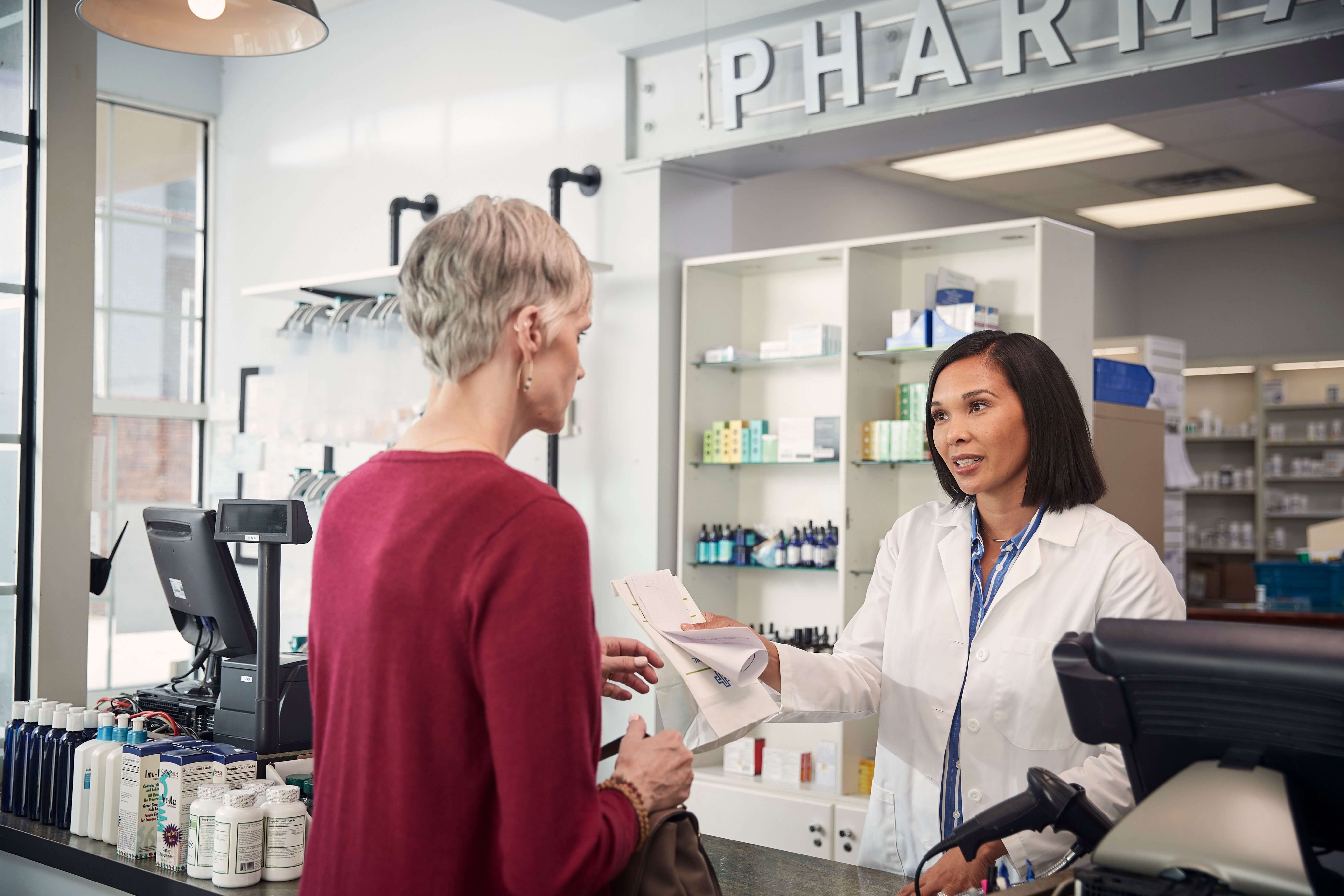 A pharmacist in a white coat assists a customer at the counter of a pharmacy, handing over medication and paperwork.