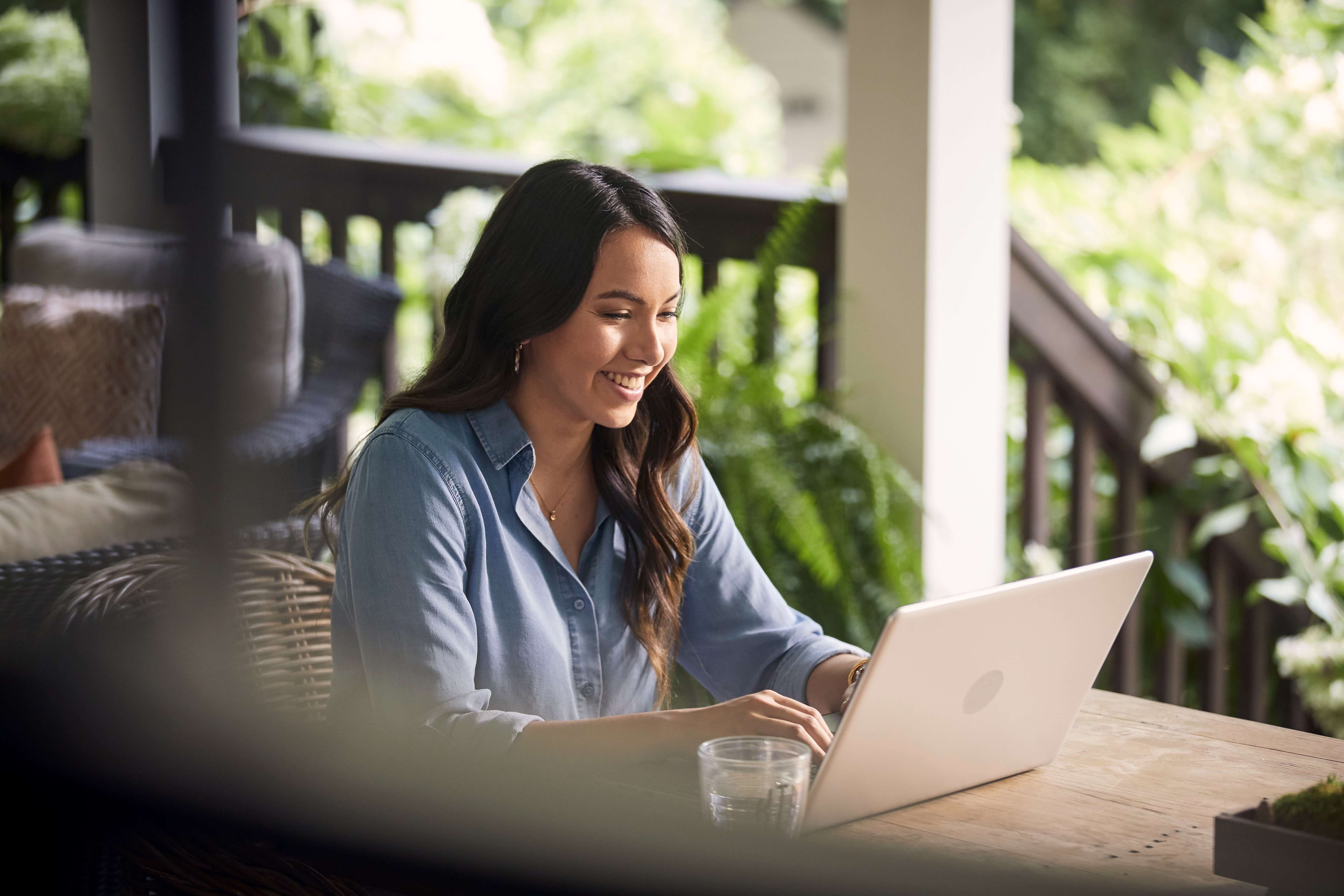 Person sitting at a wooden table on a porch, using a laptop, with greenery in the background.