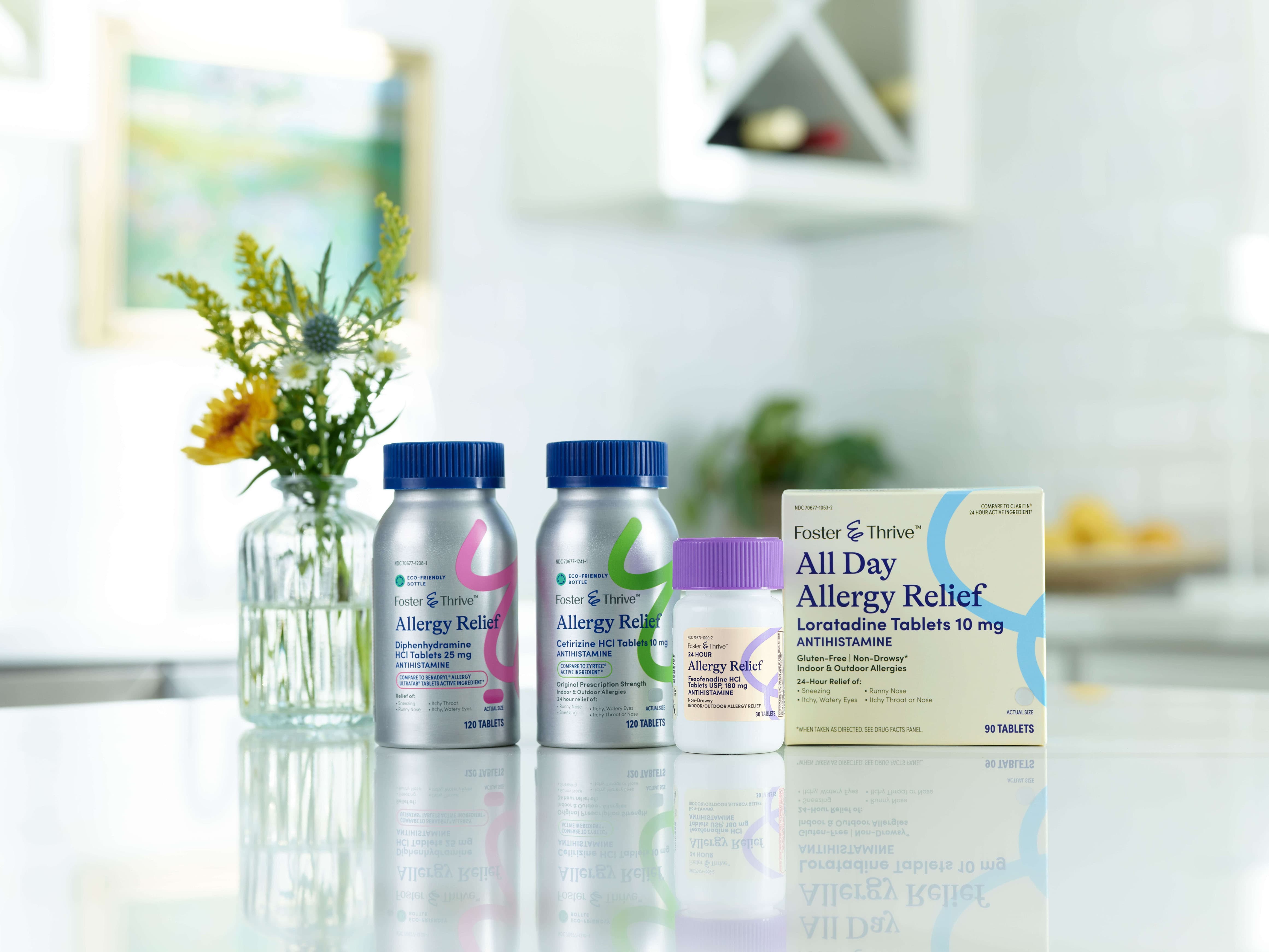 Three bottles of allergy relief medication and a box of Loratadine Tablets 10 mg on a reflective surface, with a vase of flowers in the background.