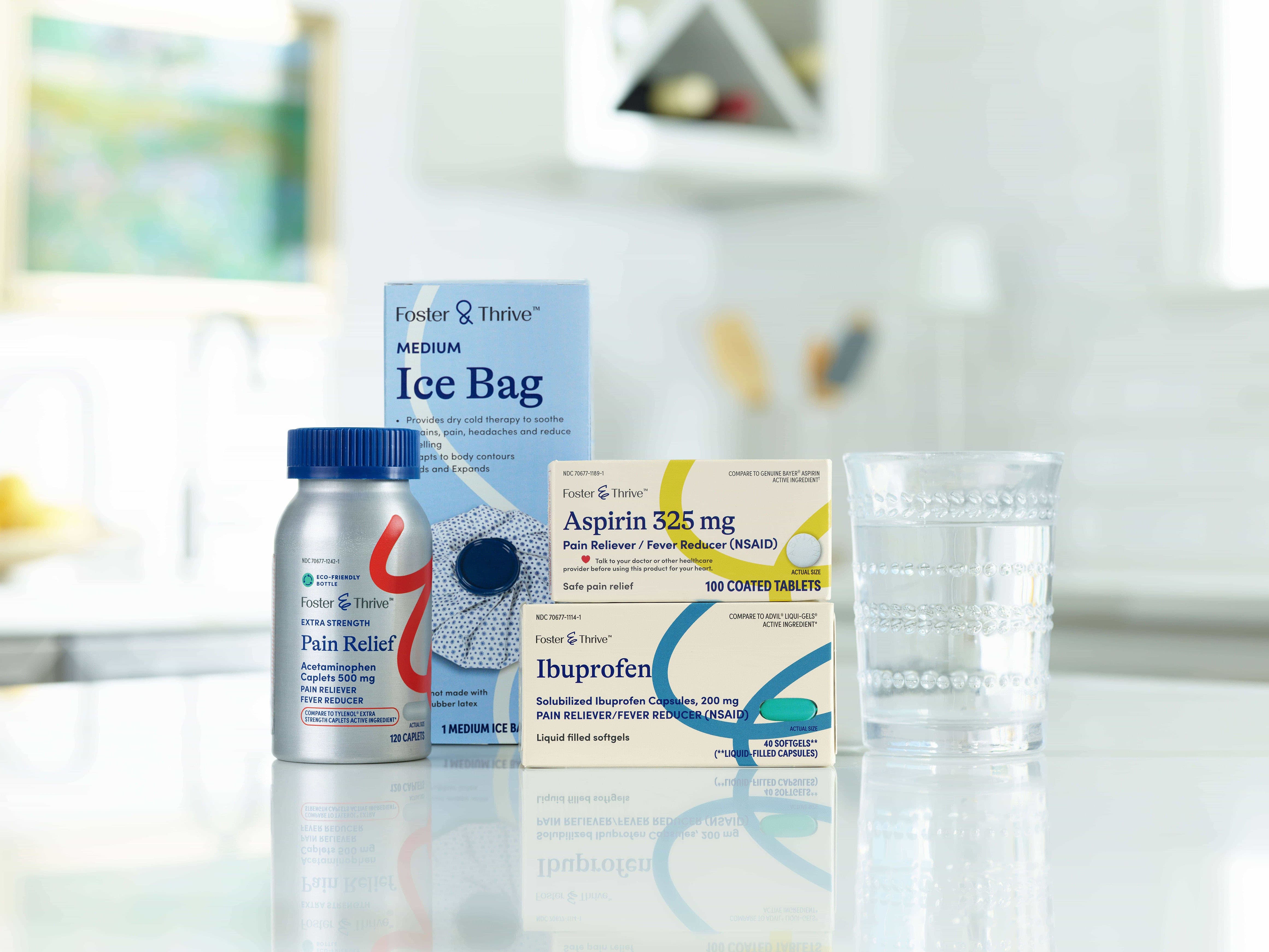 A bottle of pain relief tablets, an ice bag box, a box of aspirin, a box of ibuprofen, and a glass of water on a kitchen counter.