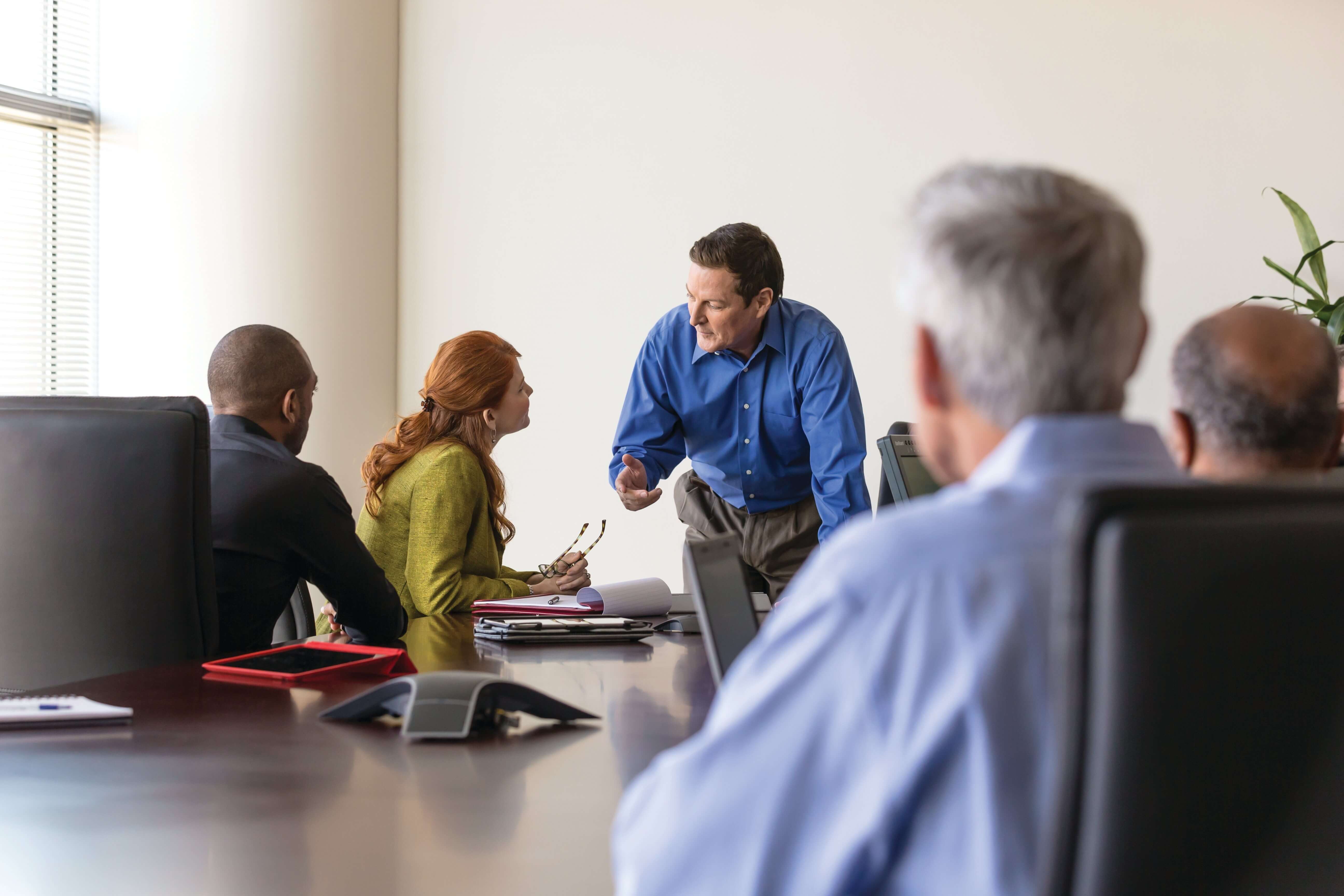 Business meeting with a man in a blue shirt discussing with colleagues around a conference table, documents and devices present.