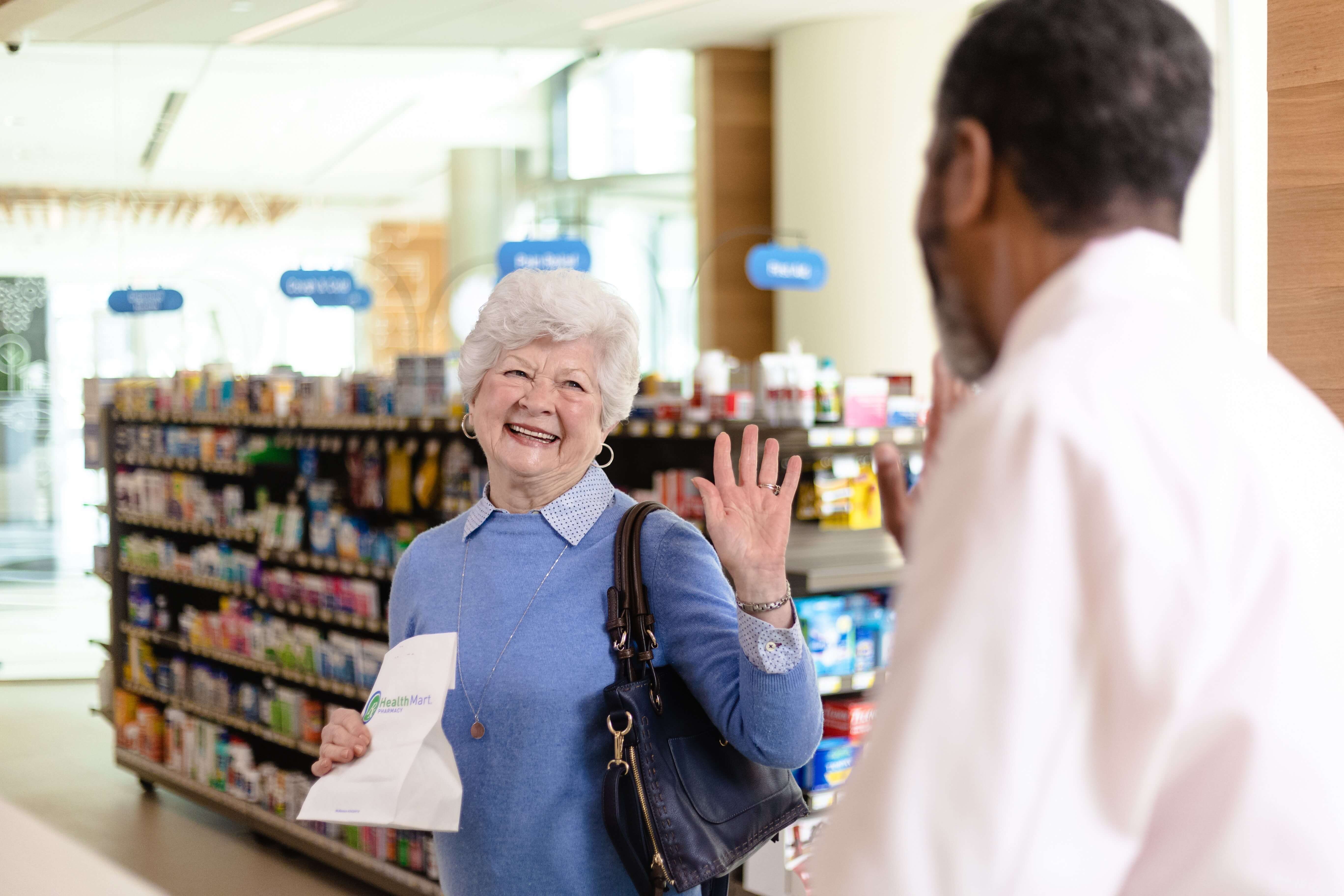 An older woman holding a pharmacy bag waves at a man in a white coat inside a store with shelves of products.