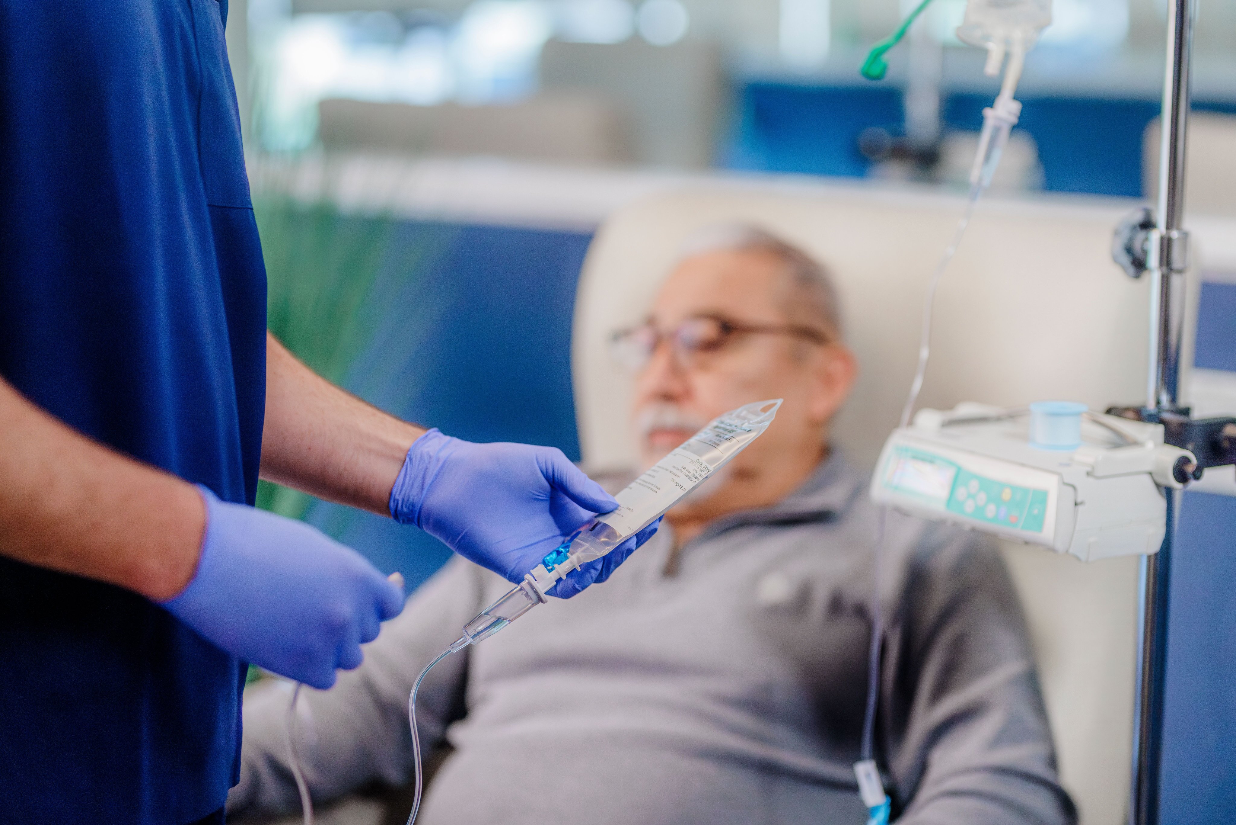 Healthcare professional in blue scrubs and purple gloves adjusting an IV line for a patient sitting in a chair.