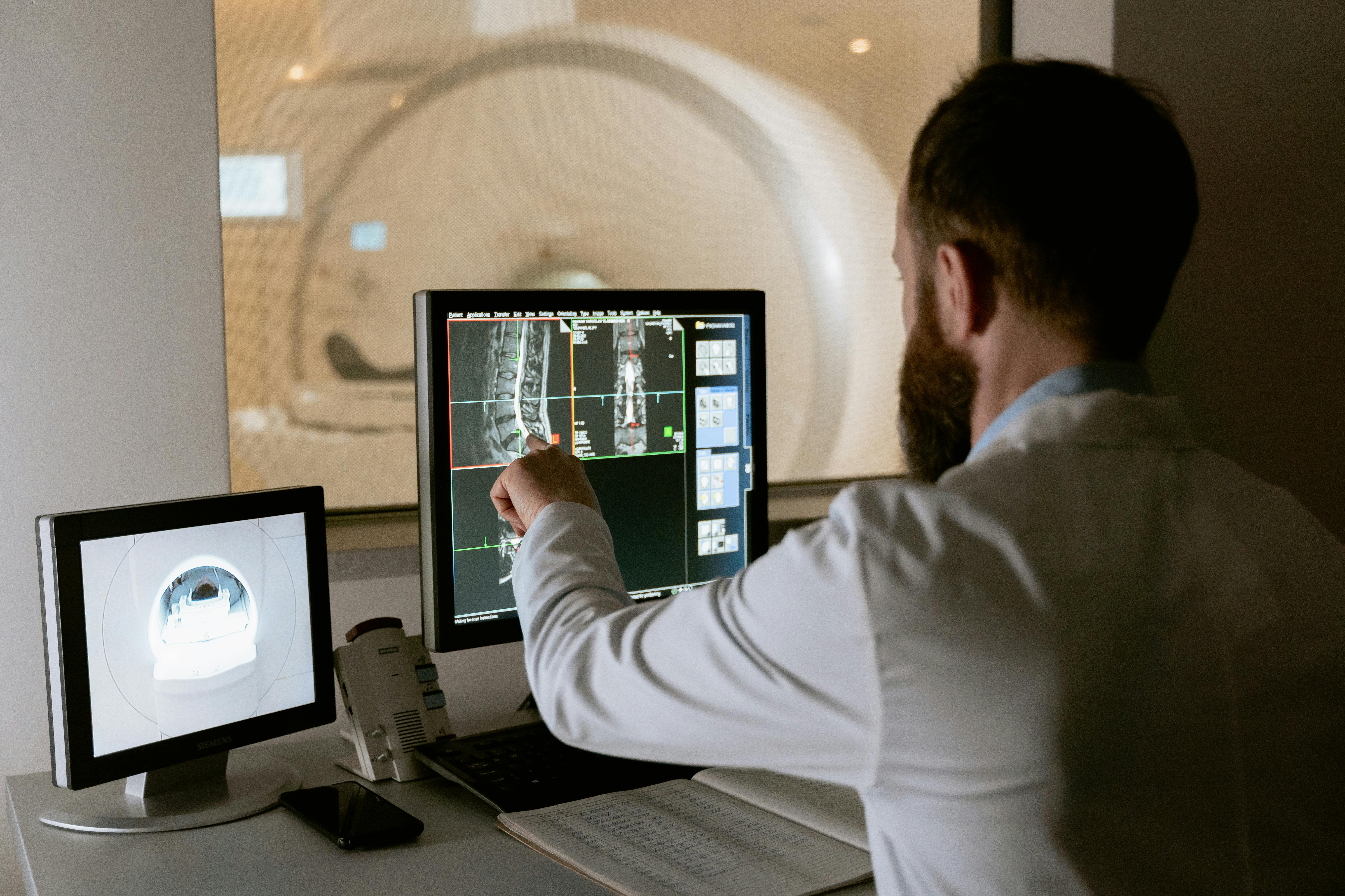 A doctor in a white coat examines MRI scans on two computer monitors.