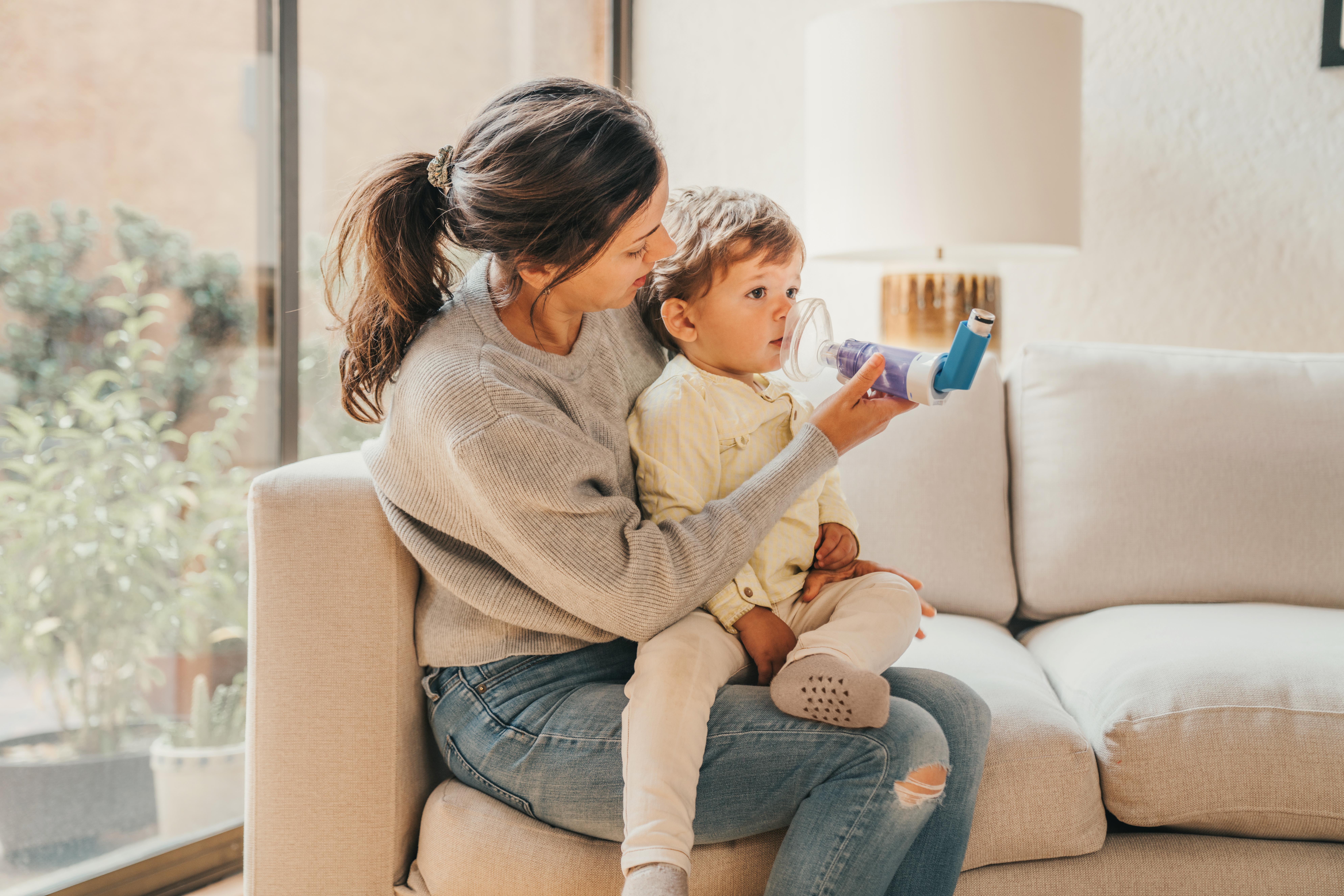 An adult sitting on a couch, holding a child who is using an inhaler with a spacer.