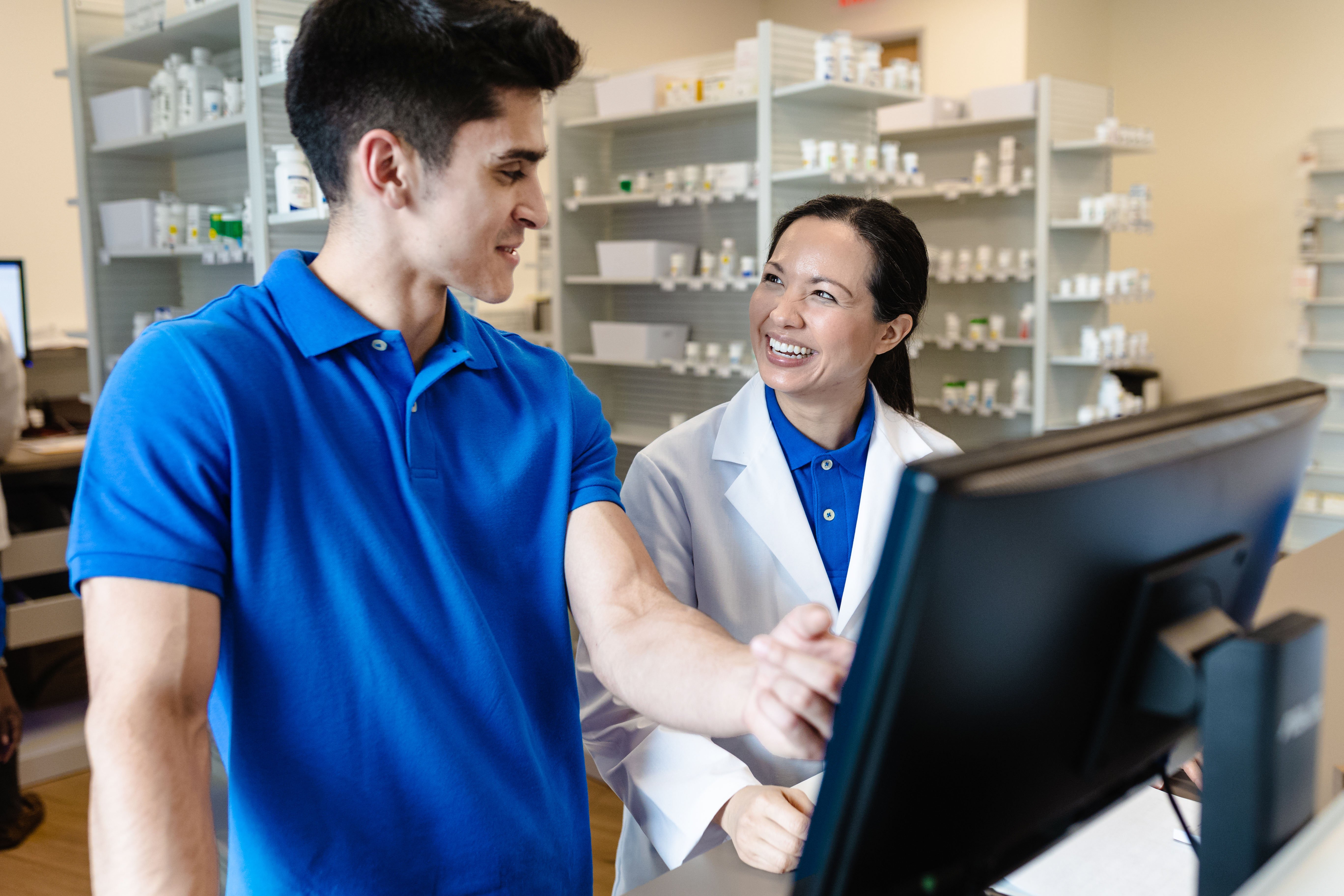A pharmacist in a white coat assists a customer in a blue shirt at a computer terminal in a pharmacy.
