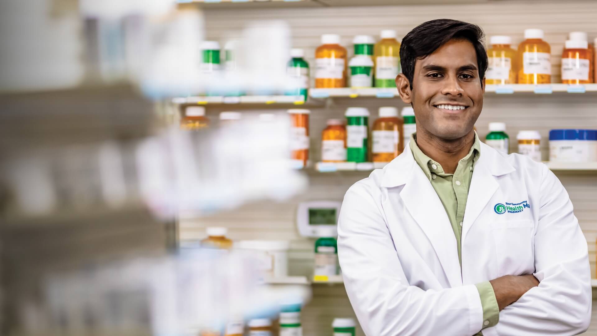 Pharmacist in a white coat stands with arms crossed in front of shelves filled with various prescription bottles.