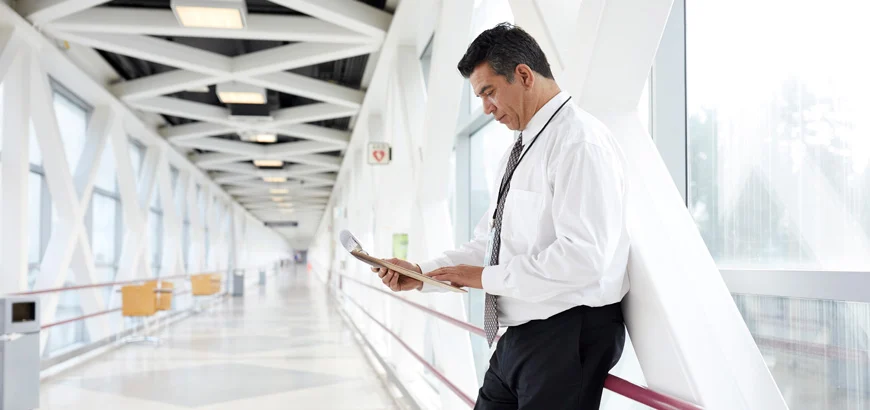 Person in a white shirt and tie reading a newspaper while leaning against the wall in a modern, bright corridor with geometric ceiling patterns.