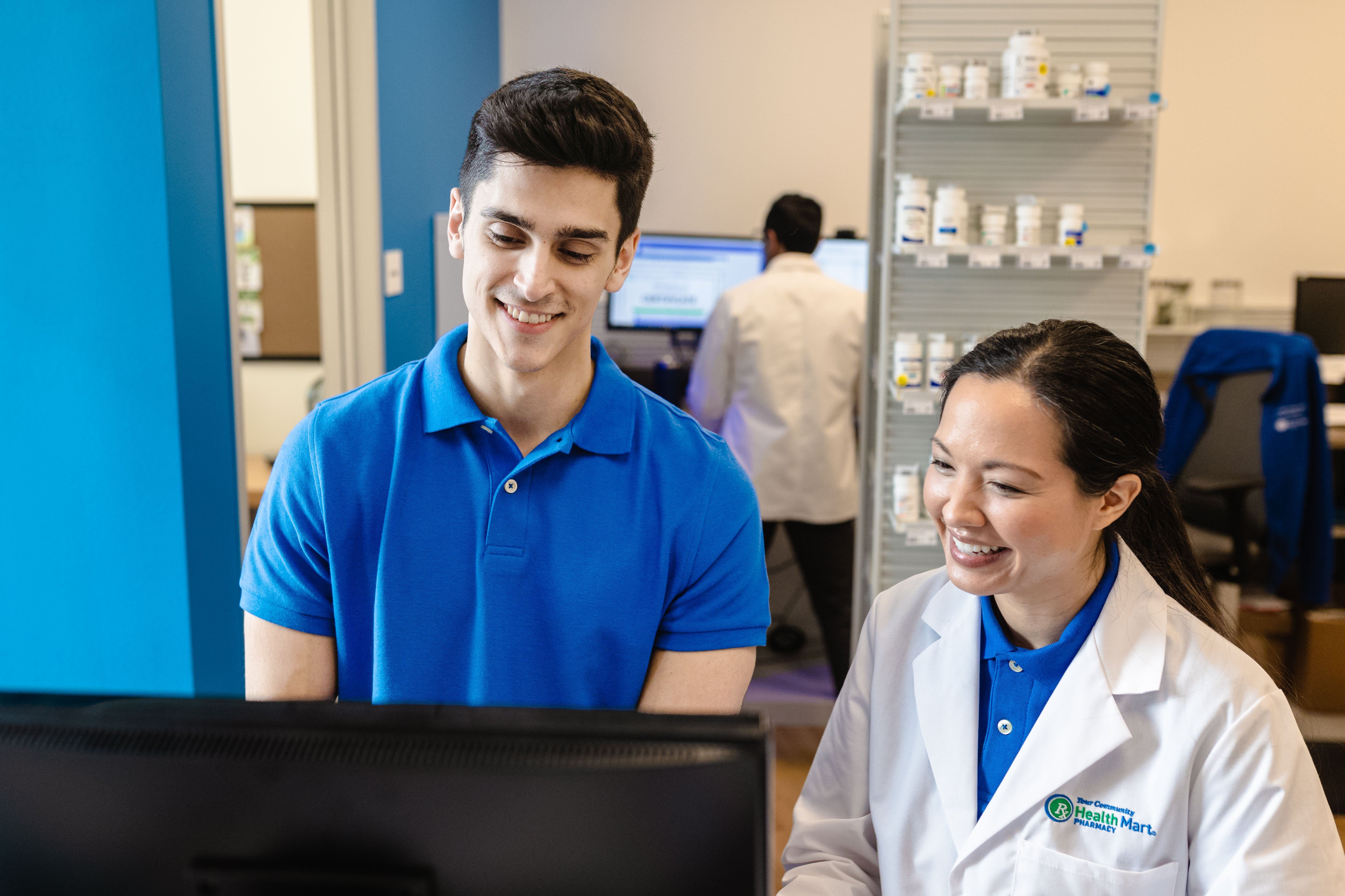 Two people in a pharmacy, one in a blue shirt and the other in a white lab coat, looking at a computer screen. Shelves with medication bottles are visible.