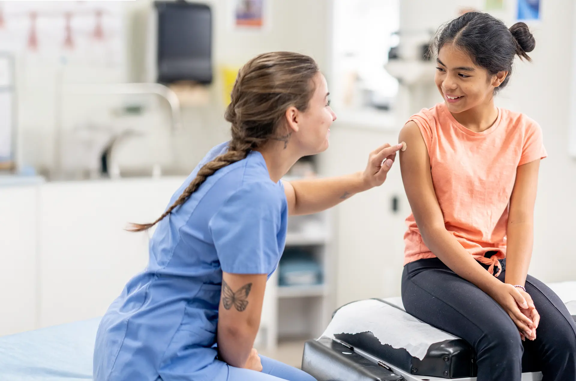 Healthcare professional in blue scrubs points to the arm of a seated patient wearing an orange shirt in a medical examination room.