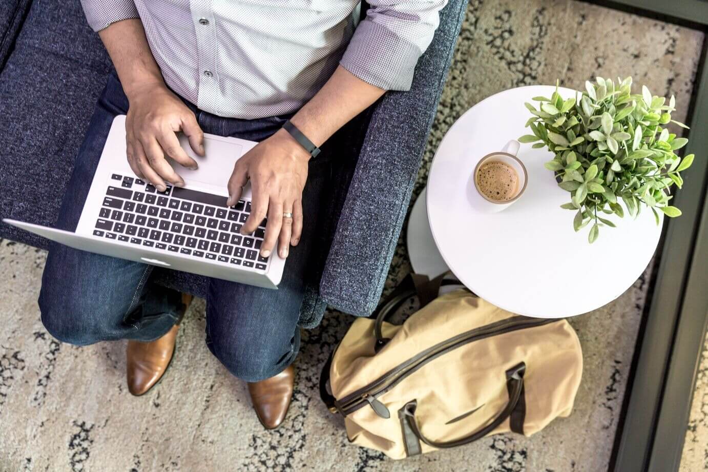 Person typing on a laptop while sitting on a couch, with a coffee cup and plant on a small table beside them, and a bag on the floor.