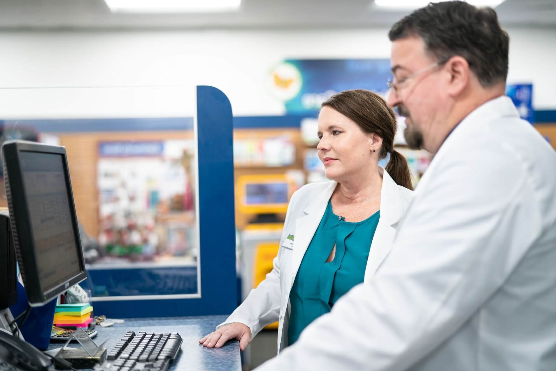 Two pharmacists in white coats working at a computer in a pharmacy.