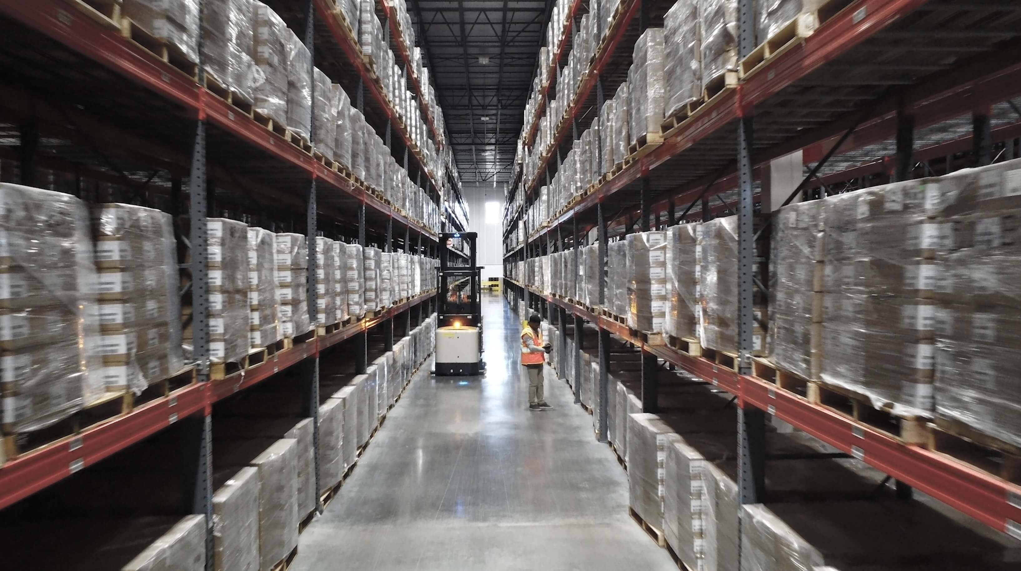 A warehouse aisle with tall shelves stacked with pallets of goods, a forklift in the middle, and a worker wearing a safety vest on the right.