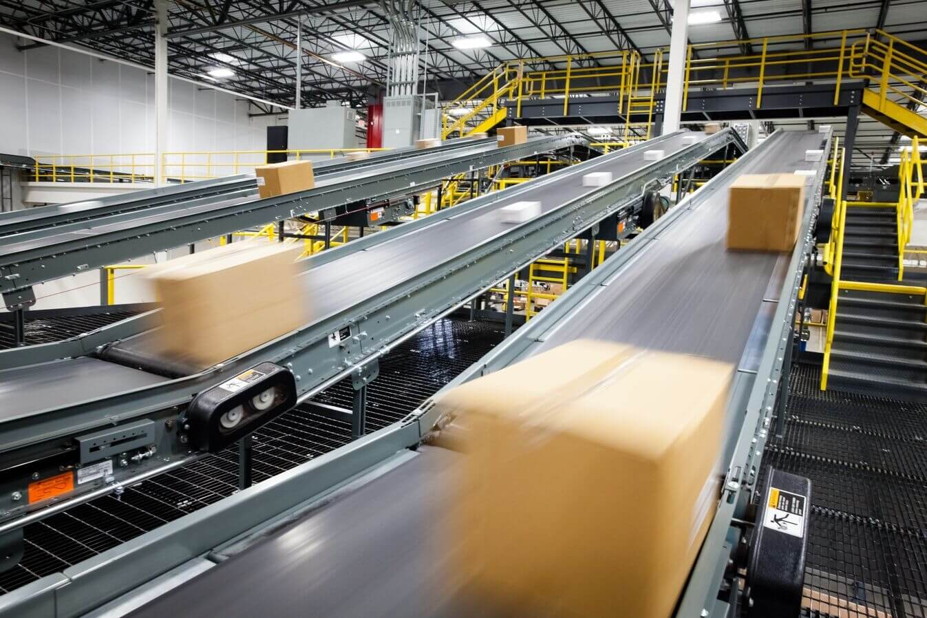 Conveyor belts with moving boxes in a large industrial warehouse with yellow railings and metal structures.