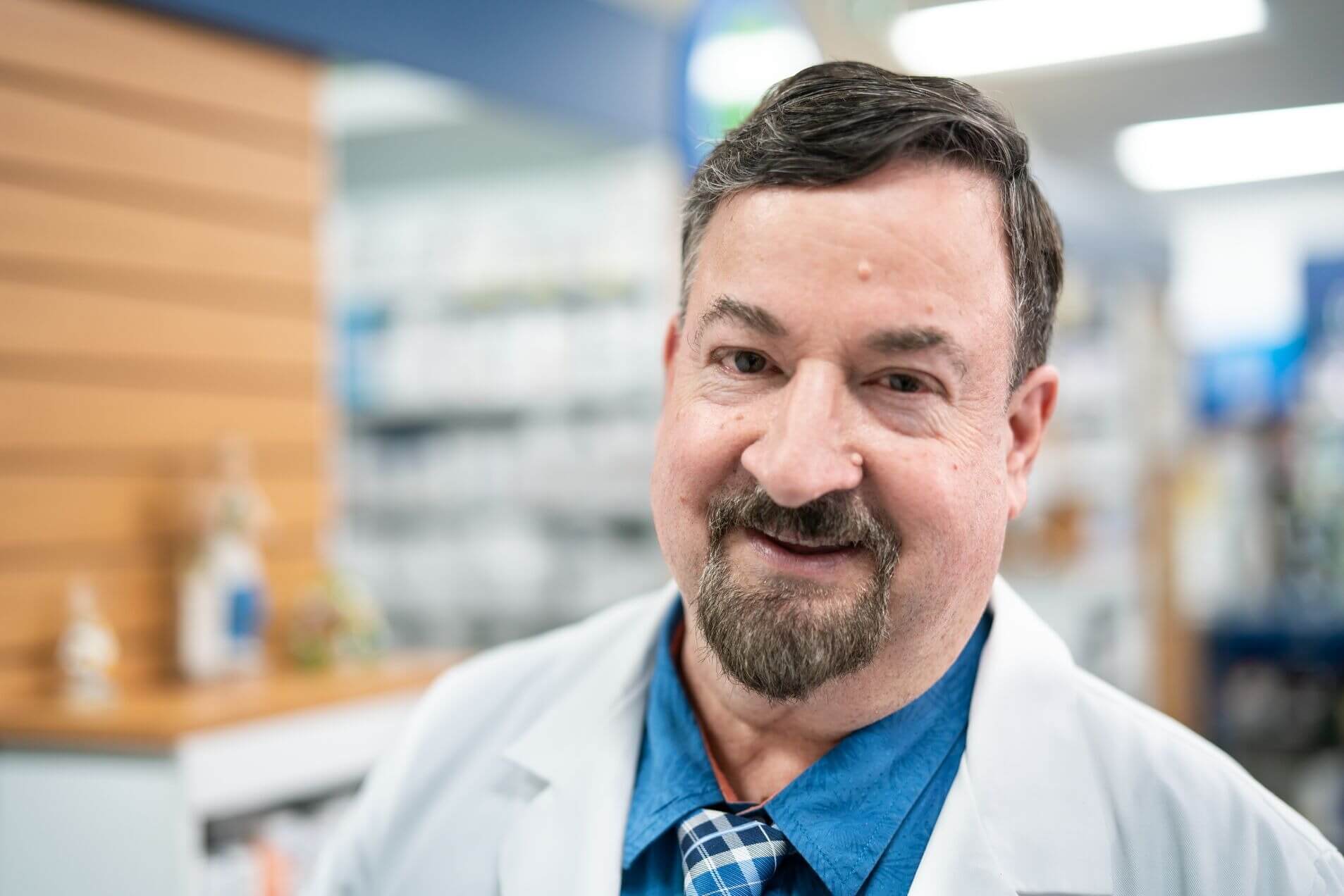 A person in a white coat stands in a pharmacy setting.
