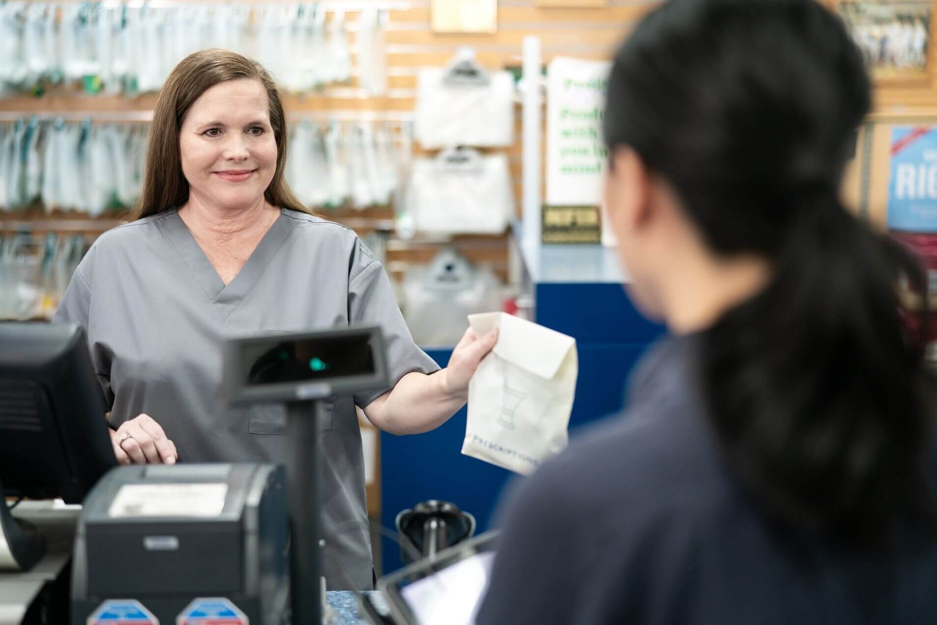 Two people in a pharmacy, one behind the counter handing a small paper bag to the other.